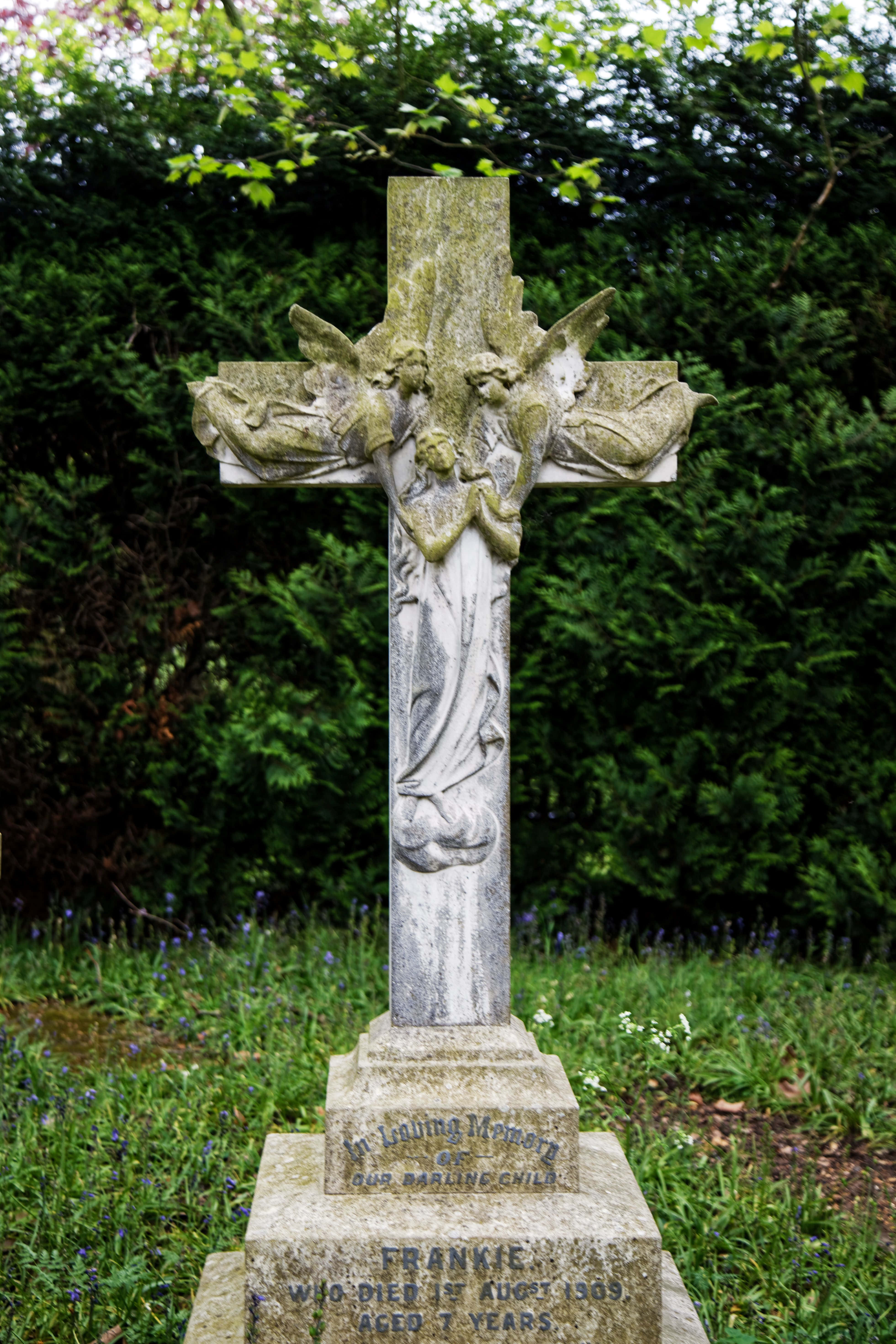 Gravestone 桌布 壁紙 — 風化的石十字架帶有天使雕刻，莊嚴地矗立在草地墓地中，周圍環繞著綠樹和藍鈴花。