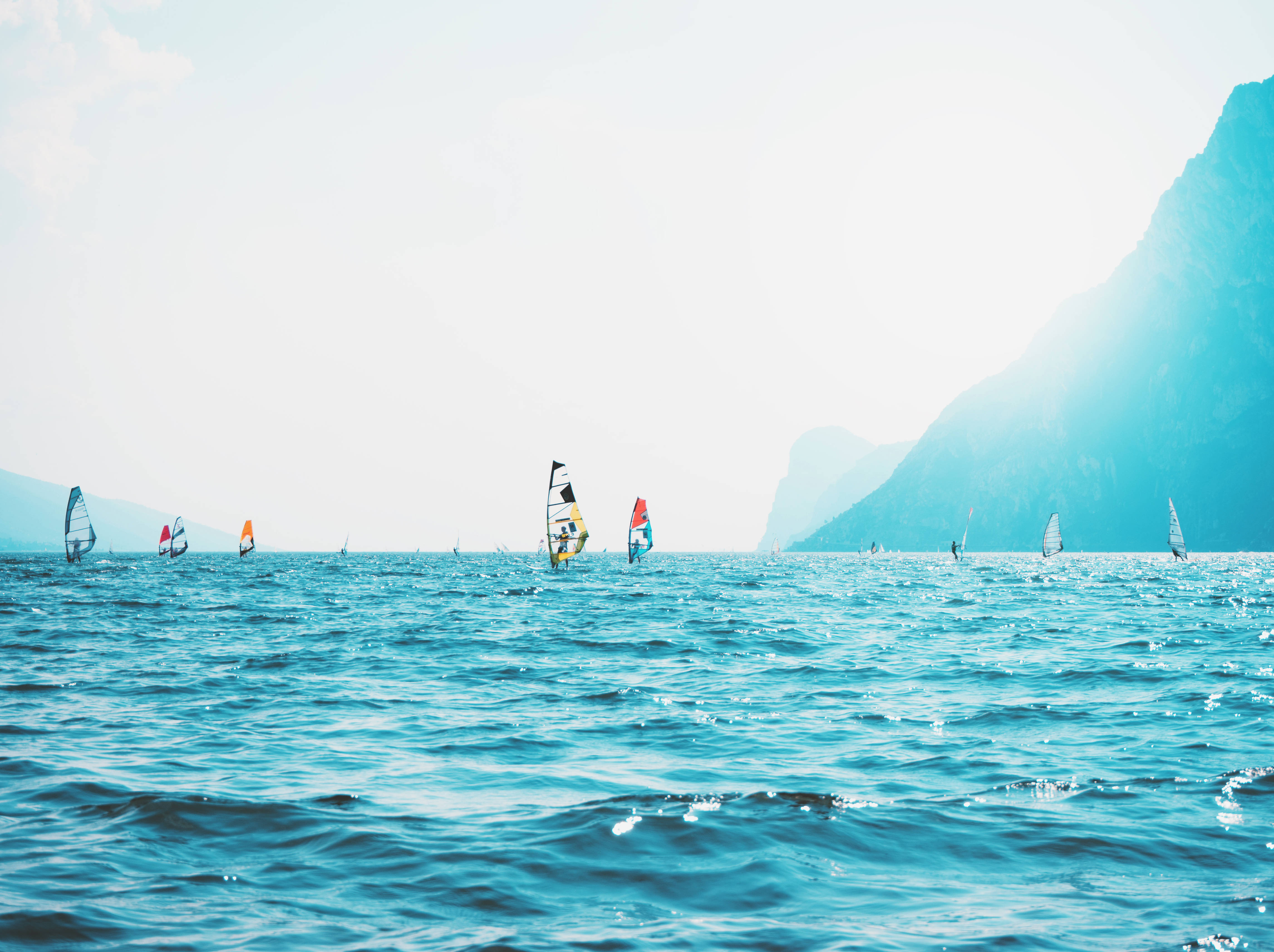 Windsurfing 桌布 壁紙 — 風帆在陽光照耀的碧綠湖面上滑行，四周是煙霧繚繞的蒼翠高山。