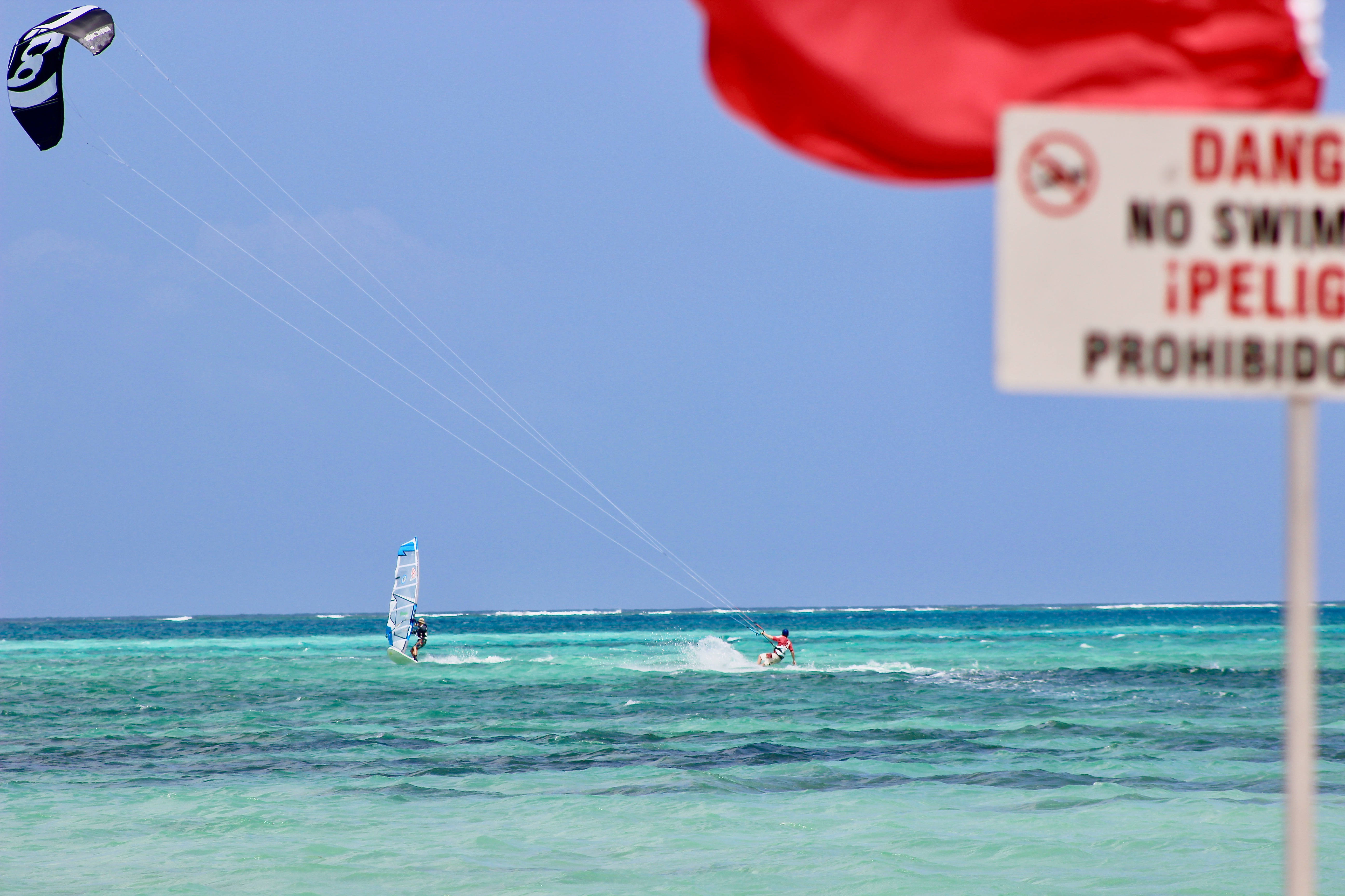 Windsurfing 桌布 壁紙 — 風帆衝浪者和風箏衝浪者在晴朗藍天下，於碧綠海水上滑行，附近有紅旗和警告標誌。