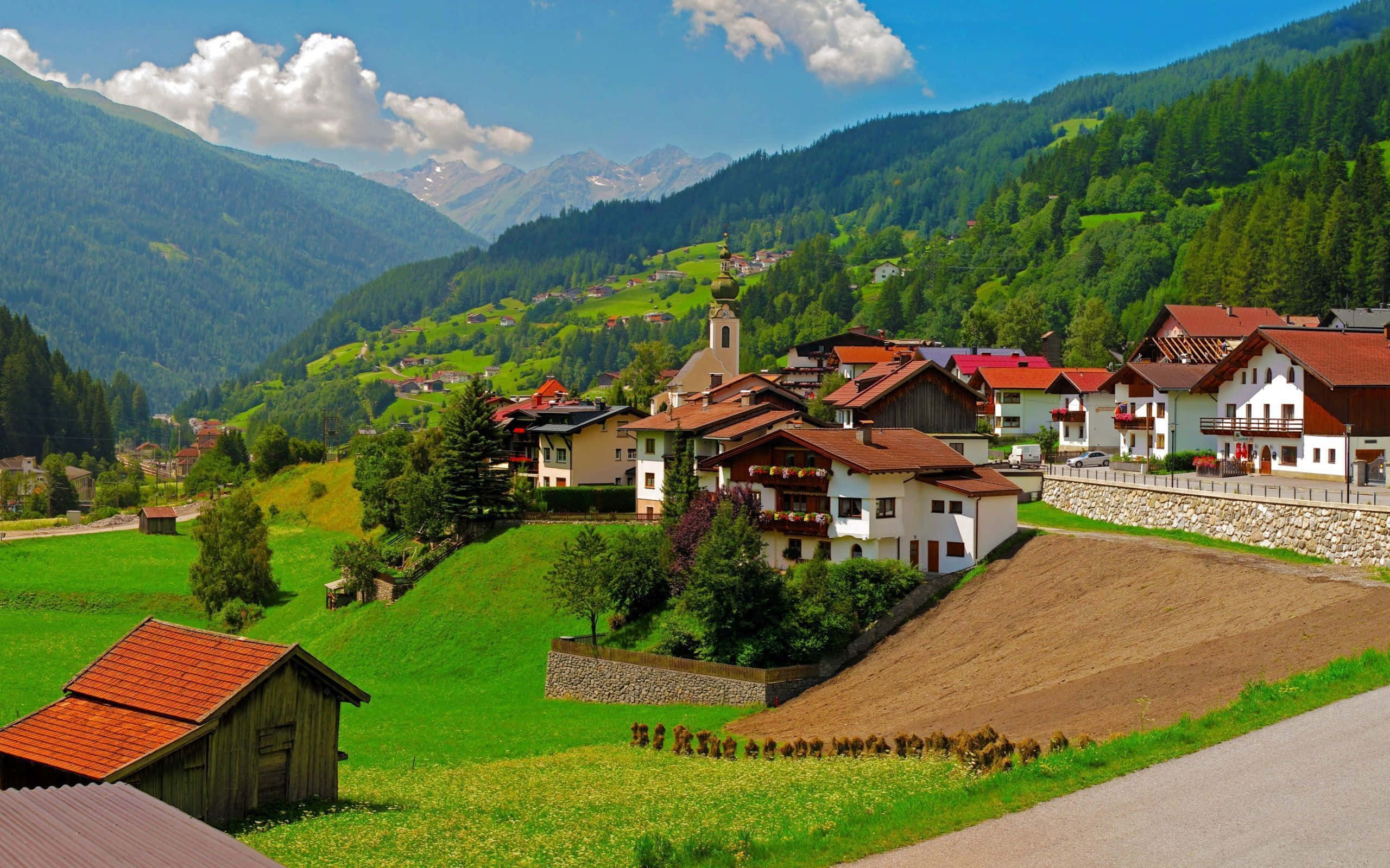 Village 桌布 壁紙 — 風景如畫的阿爾卑斯山村，坐落在鬱鬱蔥蔥的綠色山谷中，白雪皚皚的山峰在明亮的藍天下。