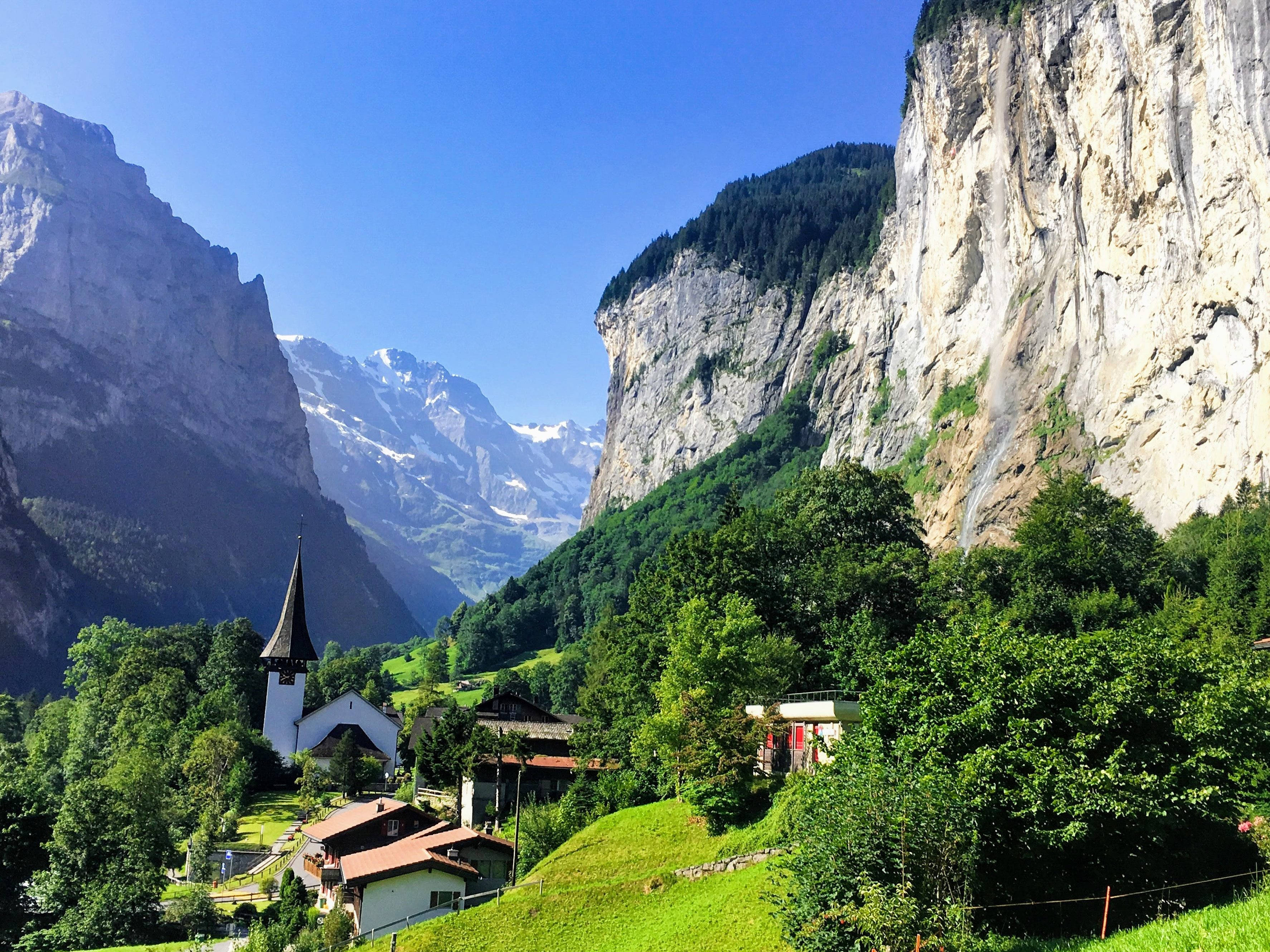 Alps 桌布 壁紙 — 風景如畫的阿爾卑斯山村，坐落於鬱鬱蔥蔥的綠色山谷中，四周是高聳的岩石峭壁和白雪覆蓋的山峰，天空湛藍清澈。