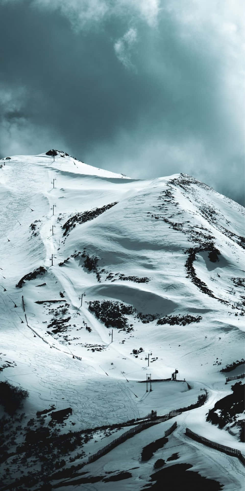  壁紙 — 風暴天空下，雪覆蓋的山坡，設有滑雪纜車和蜿蜒小徑。