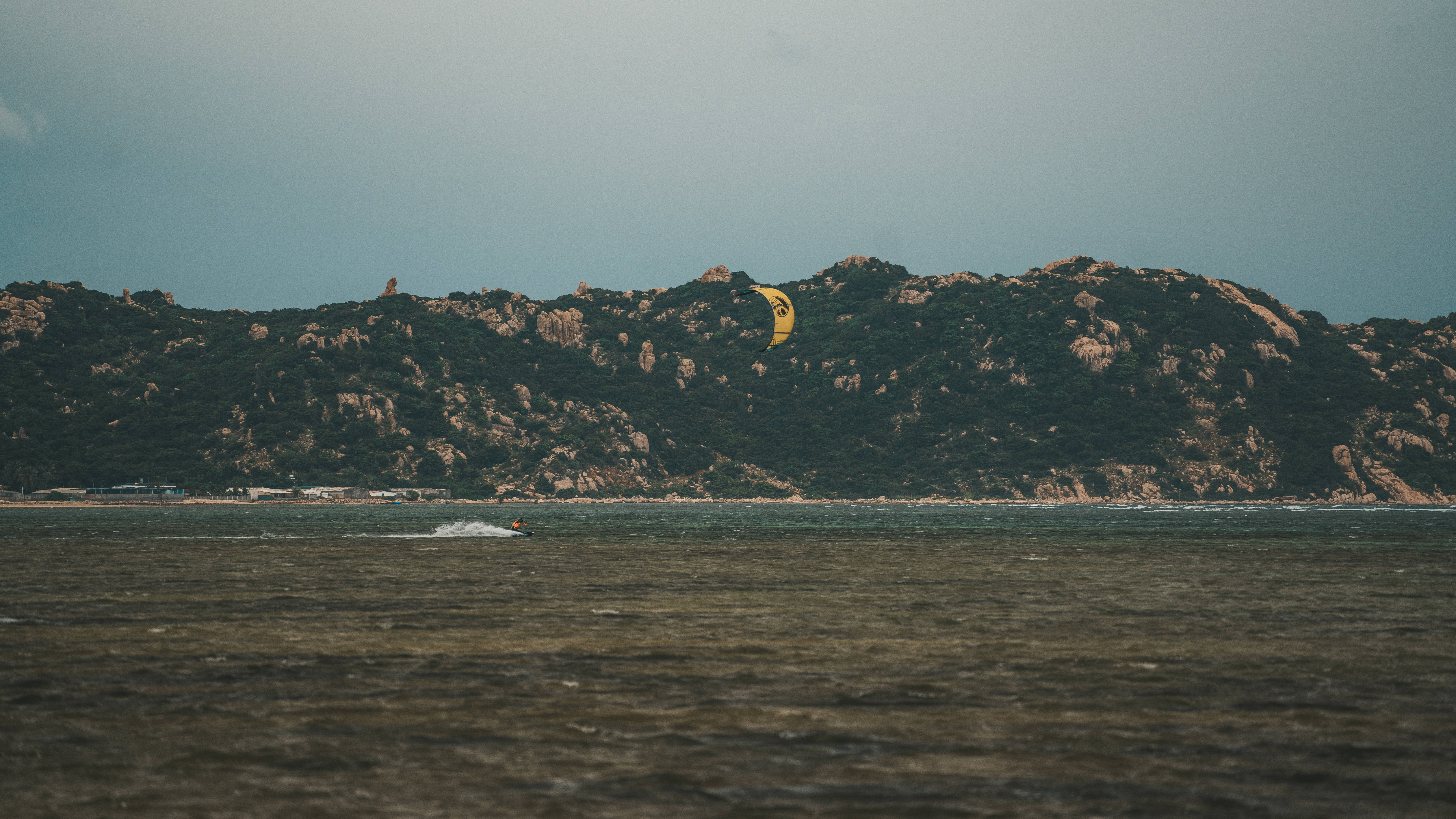 Windsurfing 桌布 壁紙 — 風箏衝浪者在平靜的水面上滑行，明亮的黃色風箏在陰天之下飛越崎嶇的綠色山丘。