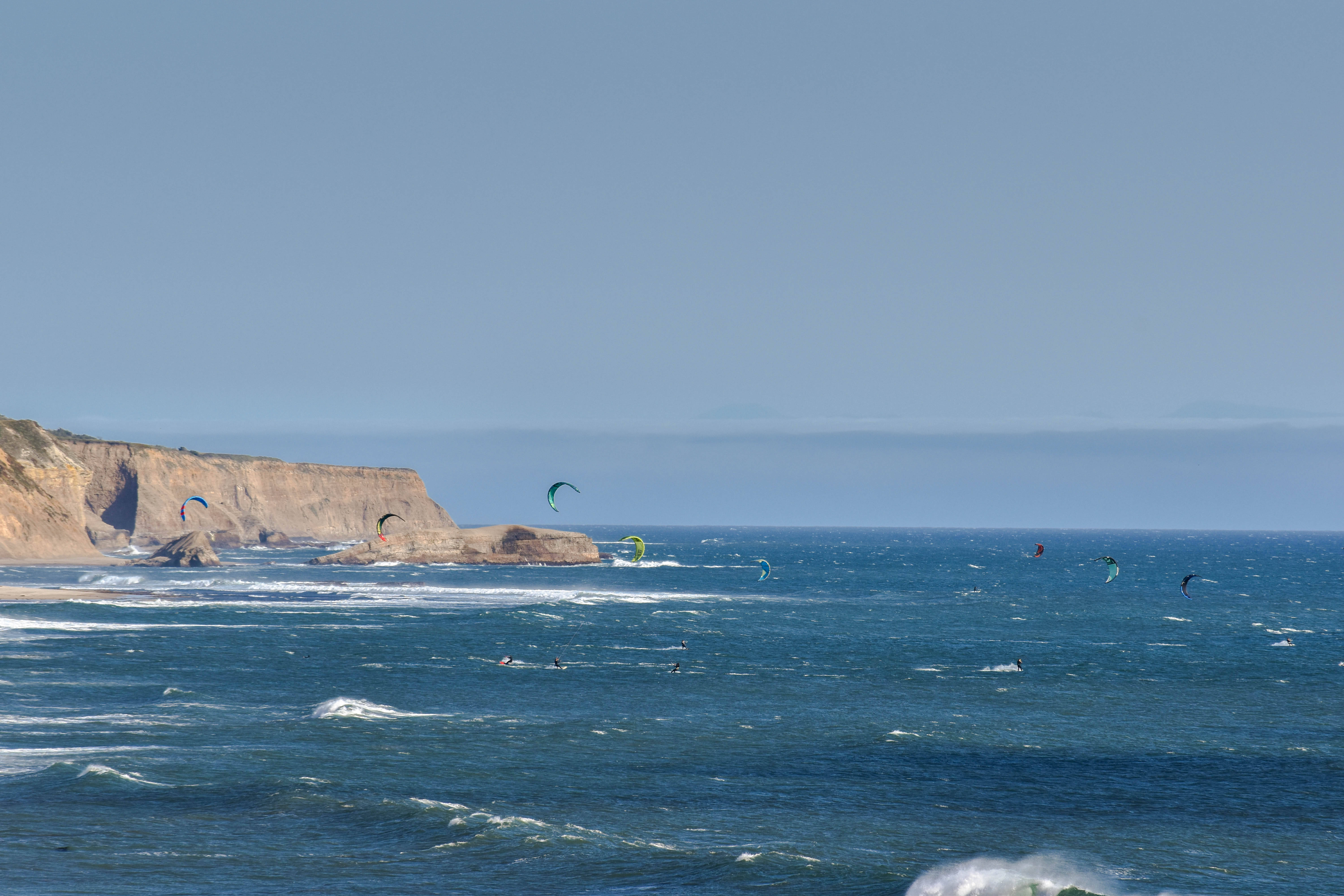 Windsurfing 桌布 壁紙 — 風箏衝浪者在晴朗天空下，於崎嶇海岸懸崖附近，滑過深藍色海浪。