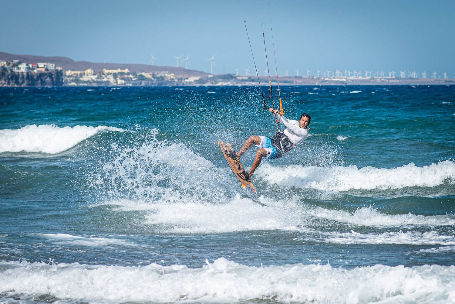 surfing 桌布 壁紙 — 風箏衝浪者在碧綠色的海浪上翱翔，遠處的懸崖上可以看到風力渦輪機，天空清澈湛藍。