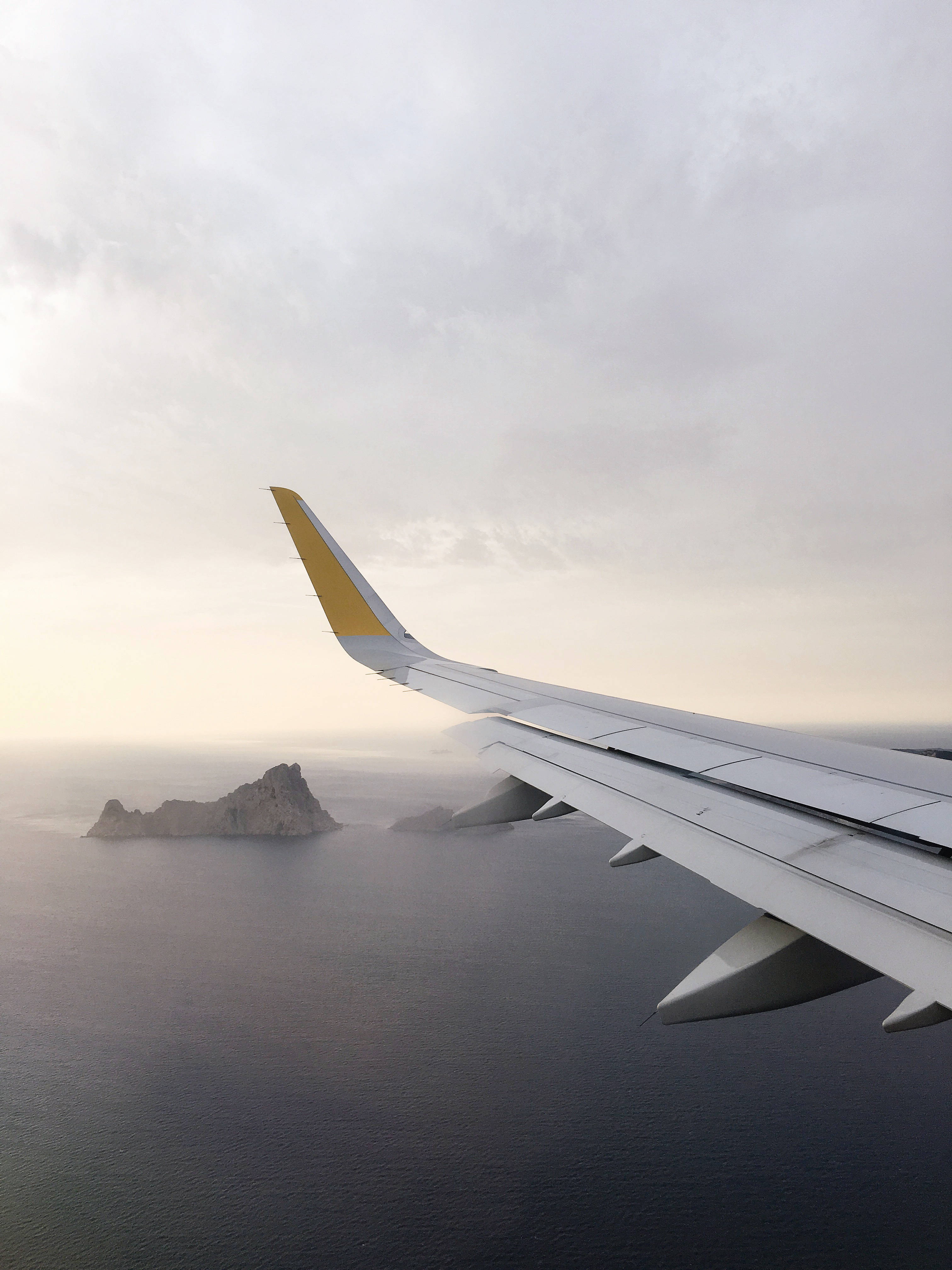 Plane 桌布 壁紙 — 飛機機翼帶黃色尖端在平靜的海洋上空滑翔，朝向岩石島嶼，下方是柔和的雲天。