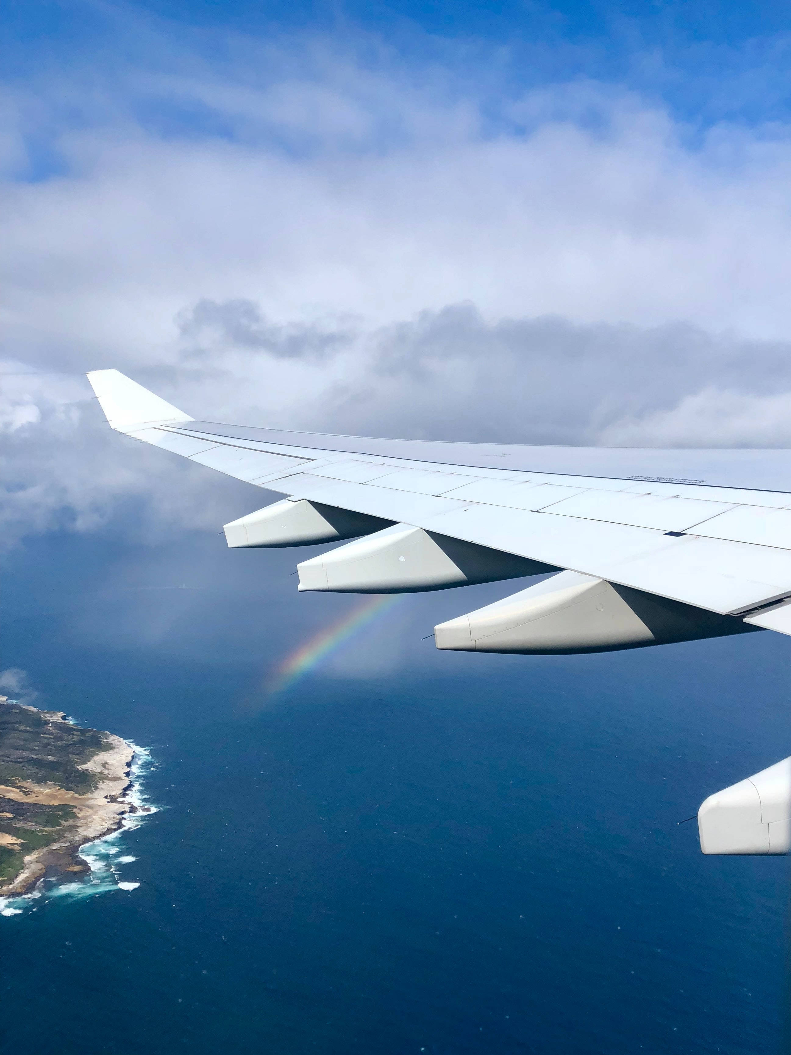 Plane 桌布 壁紙 — 飛機機翼延伸至海洋上方，一道鮮豔的彩虹在明亮的藍天下穿過雲層彎曲。
