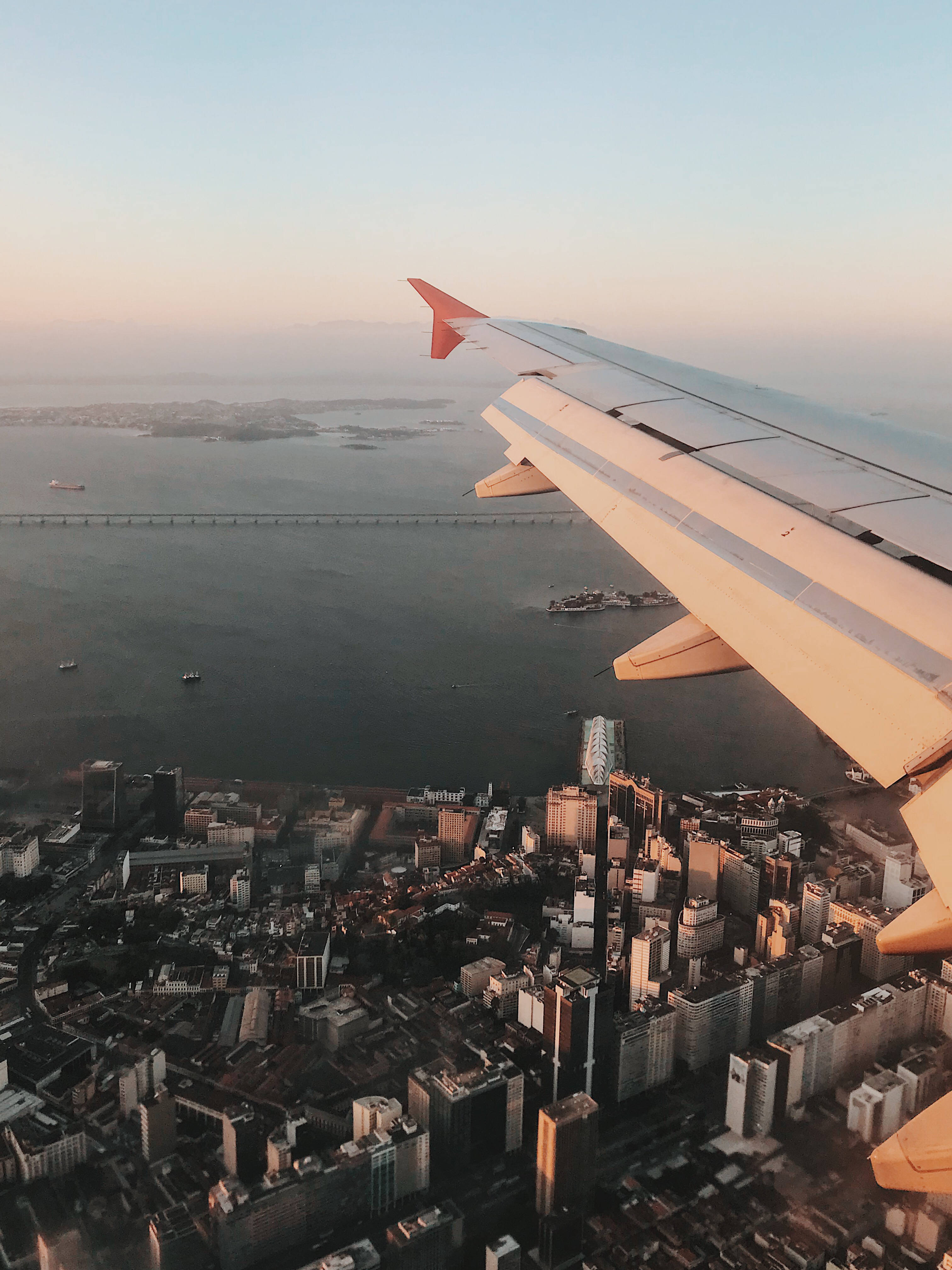 城市風景 桌布 壁紙 — 飛機翼在金色日落下滑過廣闊的海岸城市，一條長橋橫跨水面。