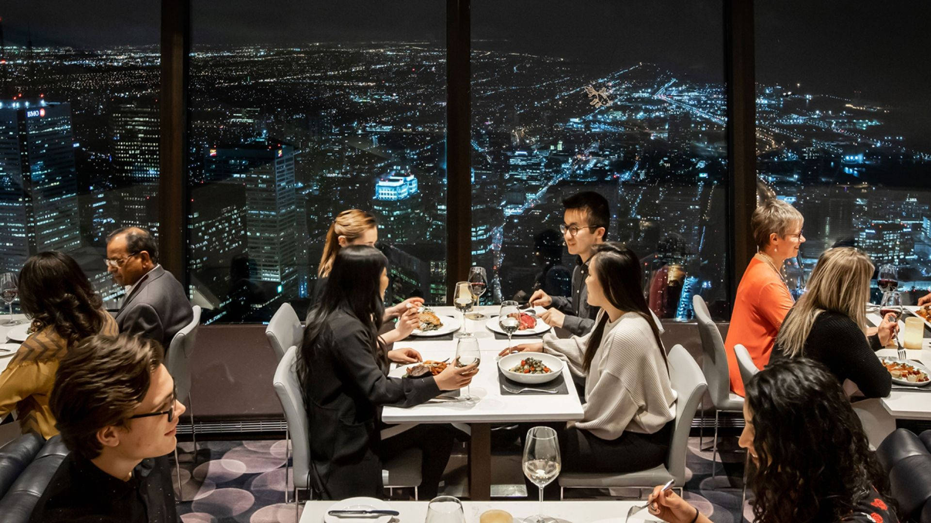 人物 桌布 壁紙 — 食客在高層餐廳享用美食，同時欣賞夜晚閃爍的城市全景。