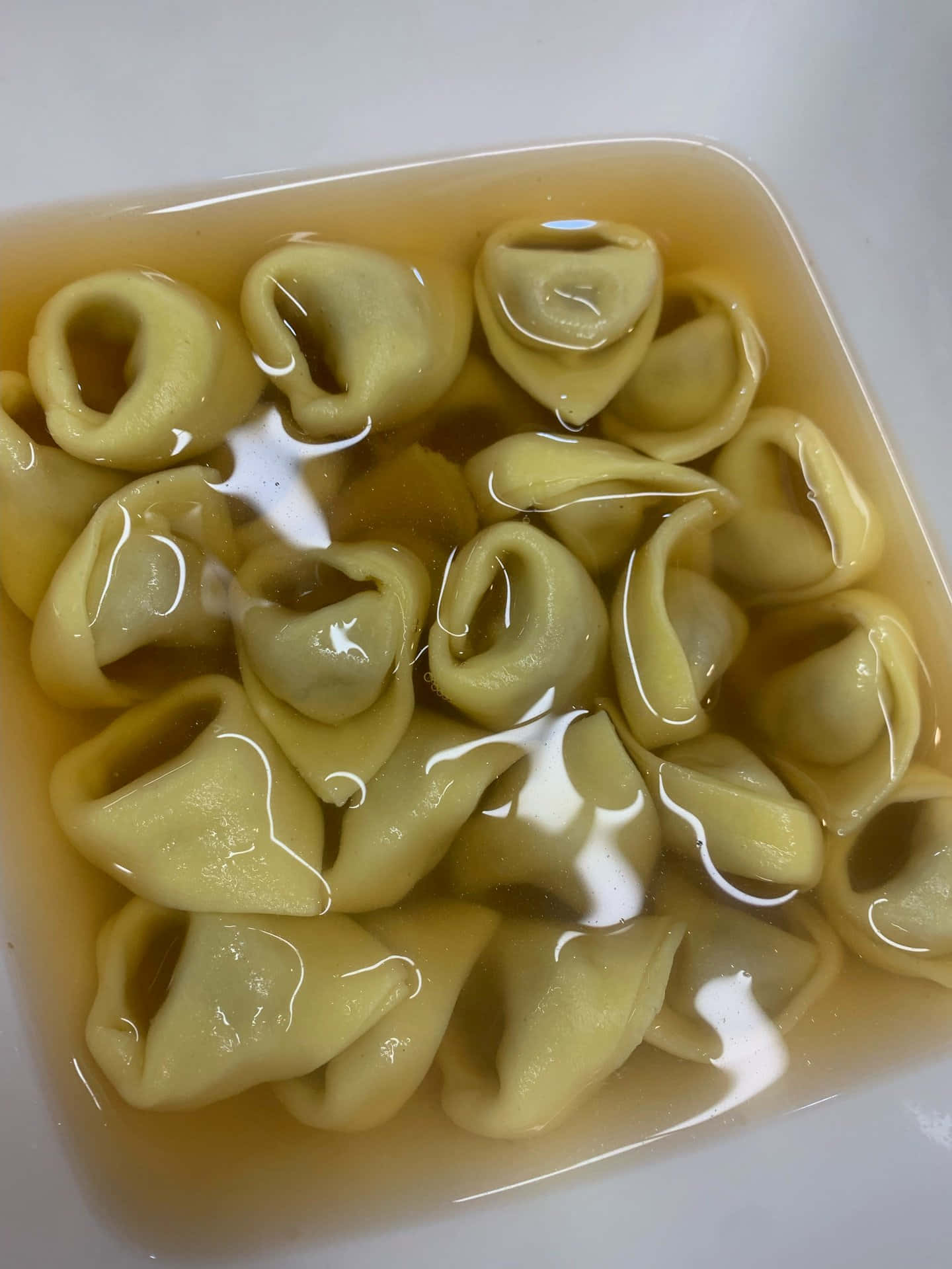 Pasta 桌布 壁紙 — 飽滿的義大利餃漂浮在金色湯汁中，置於白色方形碗內，表面有光影舞動。