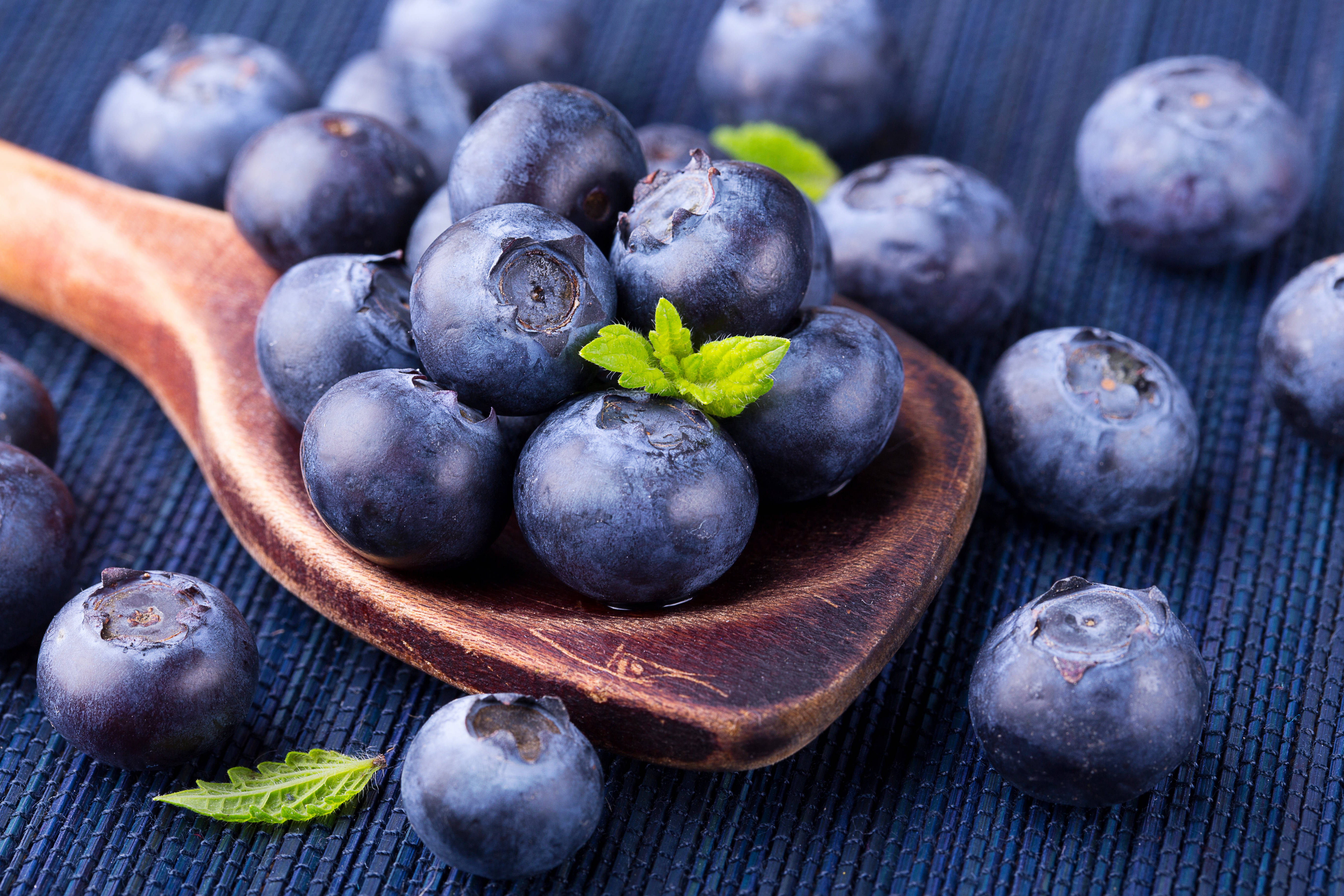 Blueberry 桌布 壁紙 — 飽滿的藍莓在木勺中閃閃發光，放在深藍色編織墊上，周圍散落著新鮮薄荷葉。
