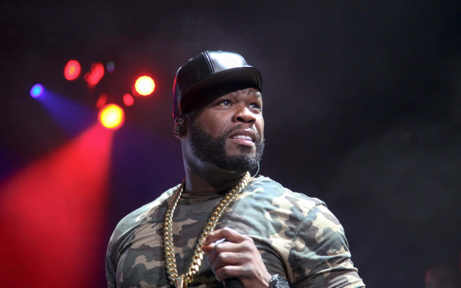 50 Cent 桌布 壁紙 — 饒舌歌手50 Cent在舞台上表演，身穿迷彩襯衫和金鏈，在彩色舞台燈光下。