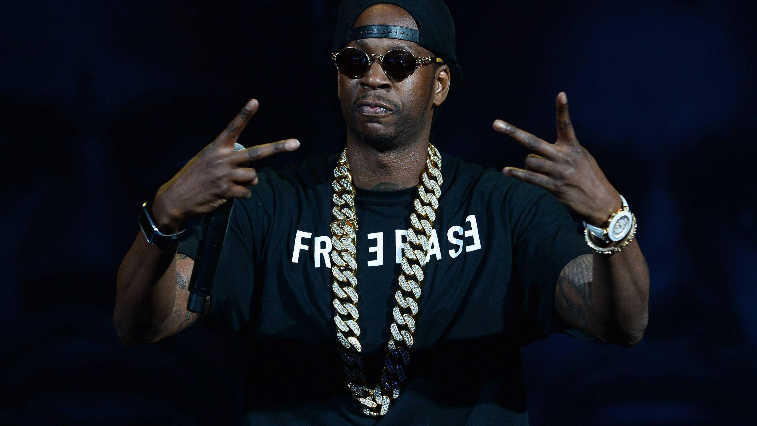 2 Chainz 桌布 壁紙 — 饒舌歌手Jay-Z在舞台上表演，戴著黑色帽子、太陽眼鏡和一條粗金鏈，同時做出手勢。