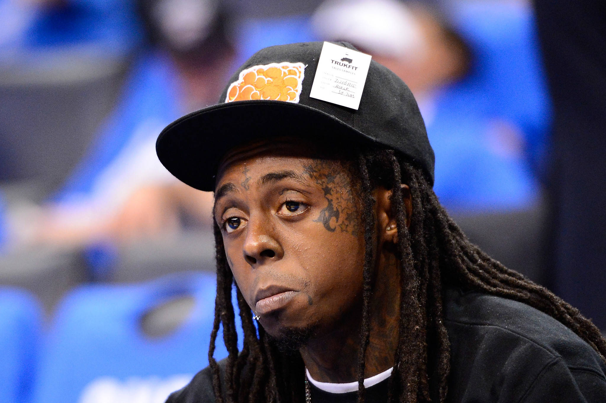 Lil Wayne 桌布 壁紙 — 饒舌歌手Lil Wayne的close-up肖像，戴著黑色帽子，上有橙色標誌和白色標籤，背景是模糊的藍色體育場座位。