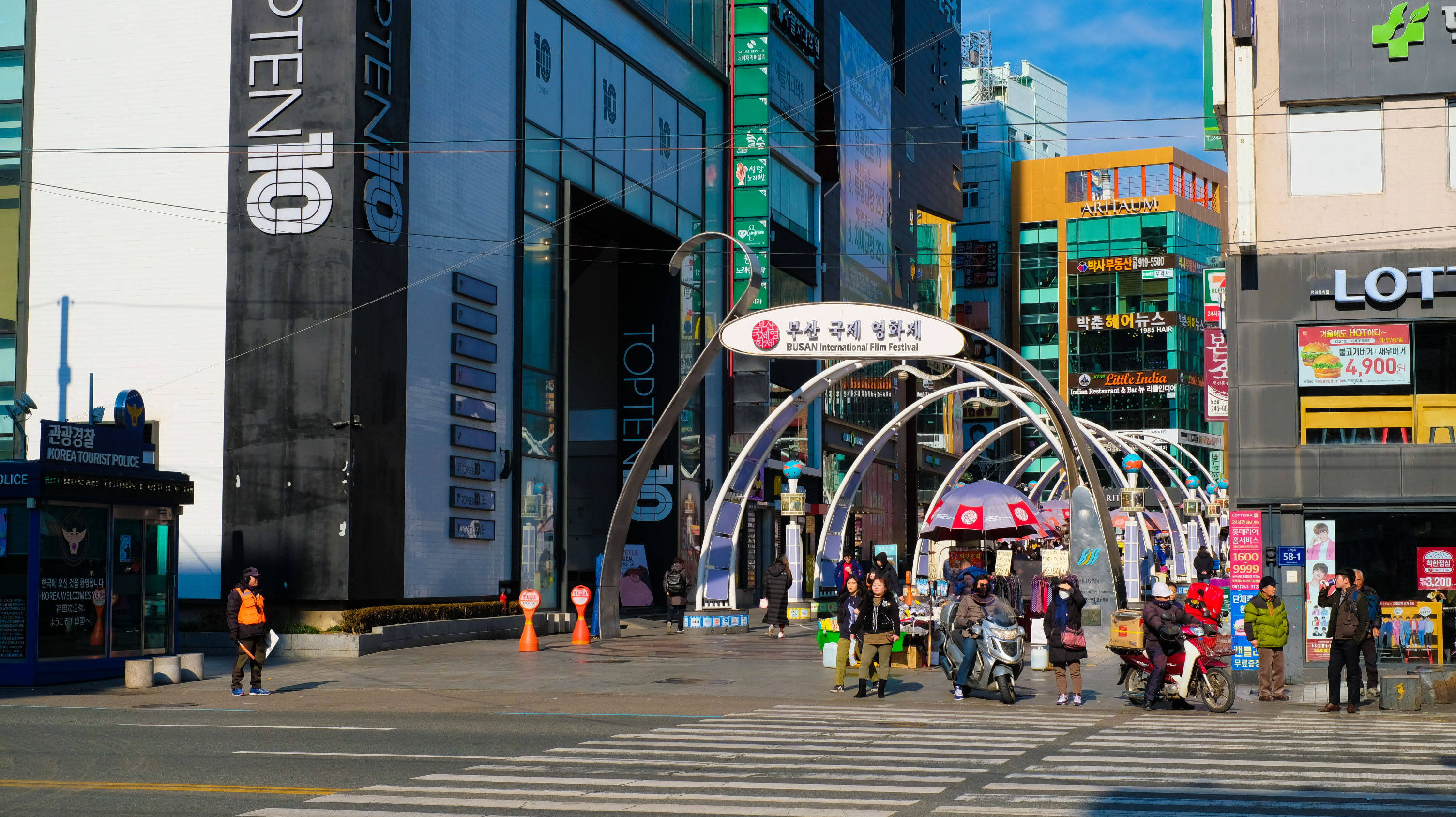 Busan 桌布 壁紙 — 首爾一個繁華的城市廣場，行人、電動滑板車和色彩繽紛的現代建築在晴朗的藍天下。