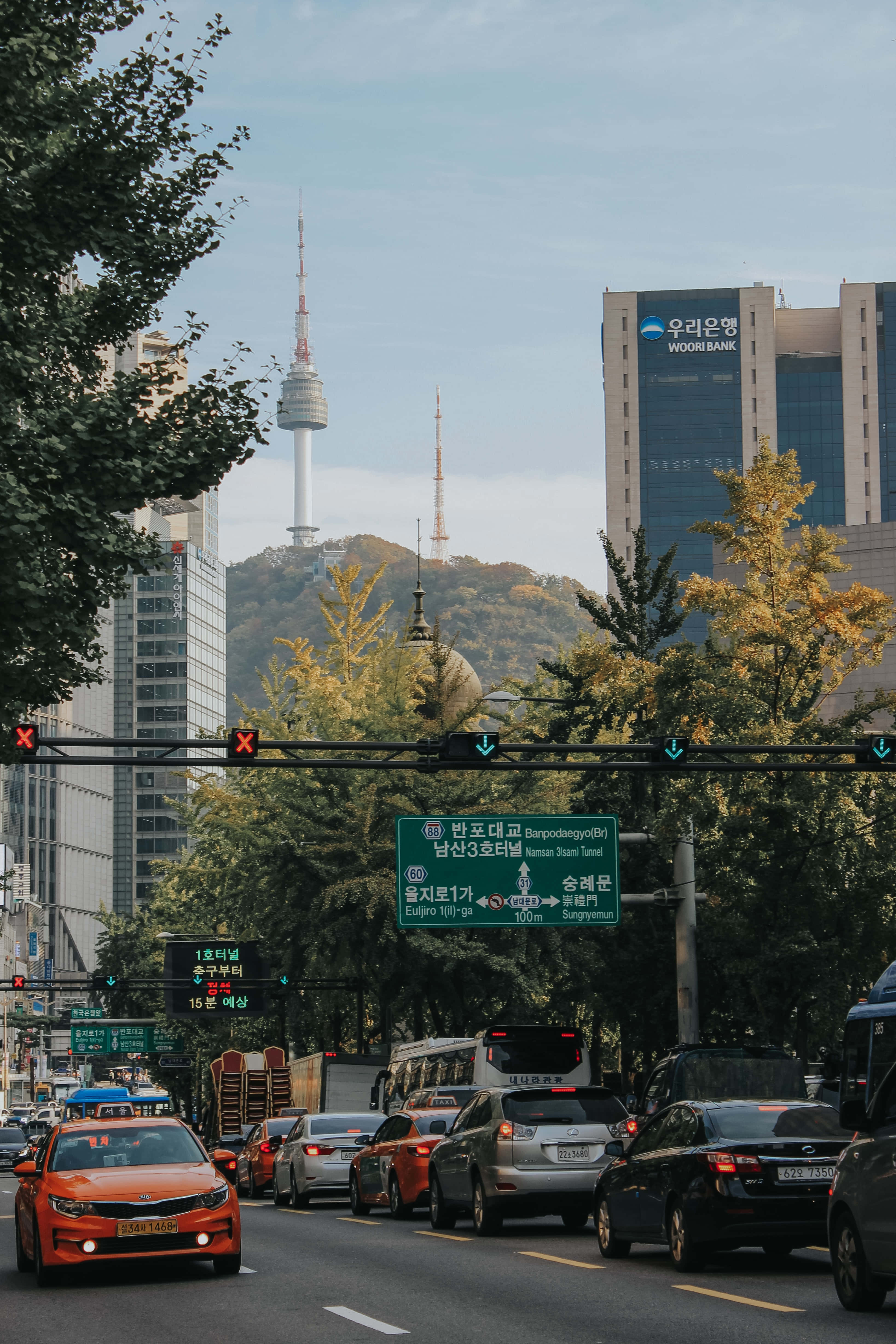 韓國 桌布 壁紙 — 首爾一條繁忙的街道，汽車在紅綠燈前停下，周圍樹木環繞，標誌性的N首爾塔在小山後方聳立。