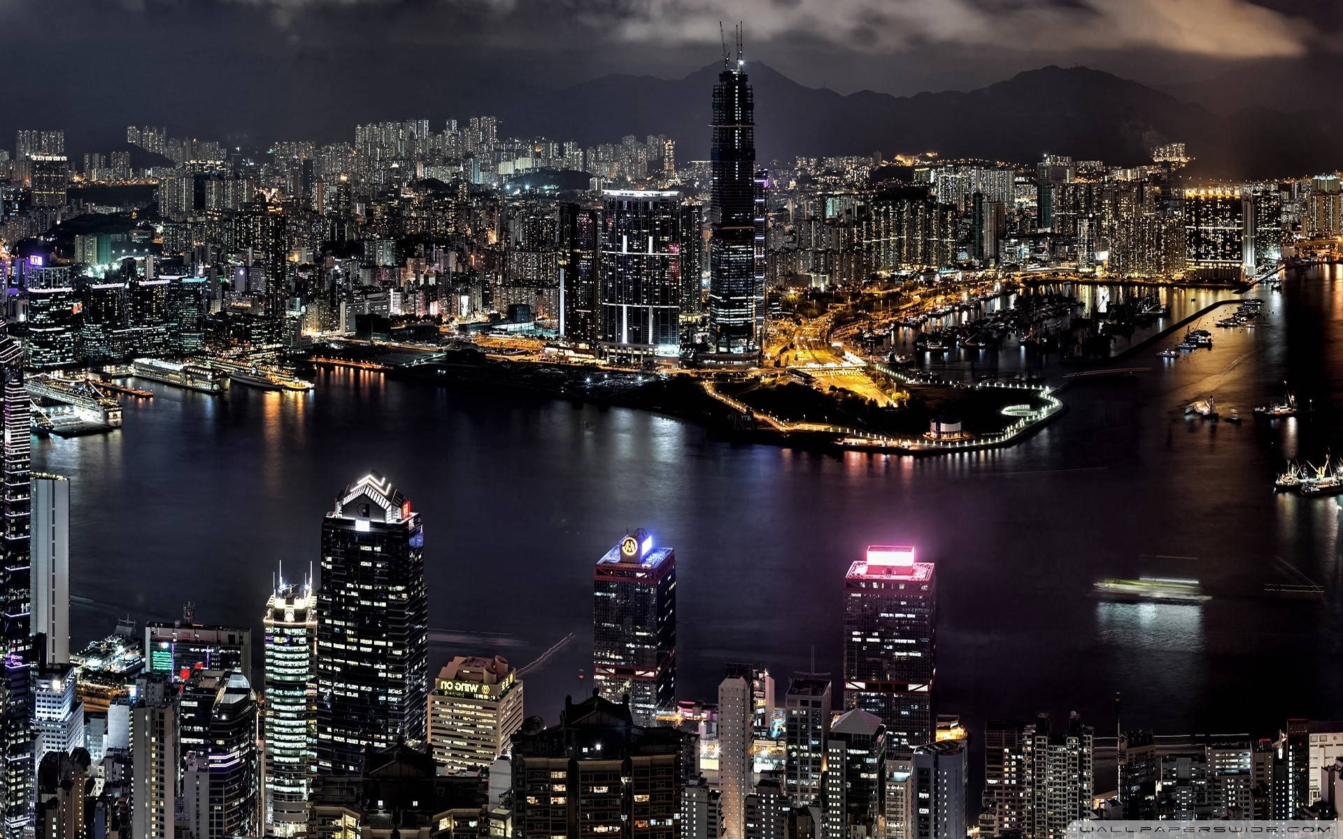 桌布 壁紙 — 香港令人眼花撩亂的夜景，高聳的摩天大樓在陰天下的黑暗海港上反射著霓虹燈光。