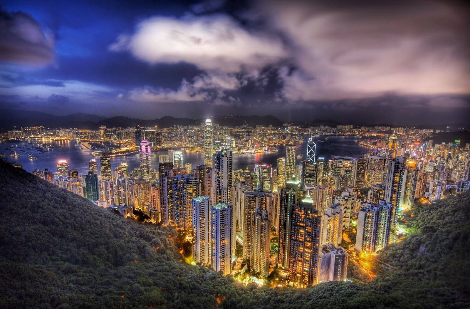 Bogota 桌布 壁紙 — 香港令人驚嘆的夜景，摩天大樓在戲劇性的多雲天空下閃閃發光，坐落在鬱鬱蔥蔥的綠色山丘之間。