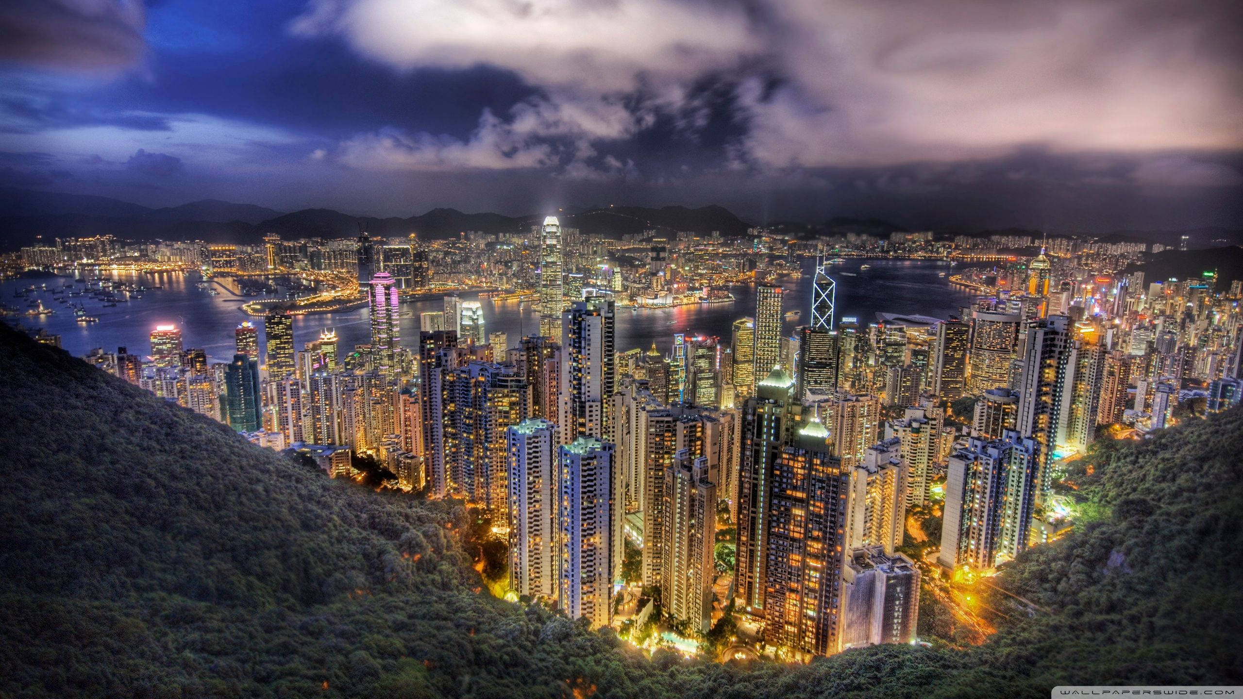城市風景 桌布 壁紙 — 香港令人驚嘆的夜景，閃爍的摩天大樓坐落在鬱鬱蔥蔥的綠色山丘之間，天空烏雲密布，極具戲劇性。