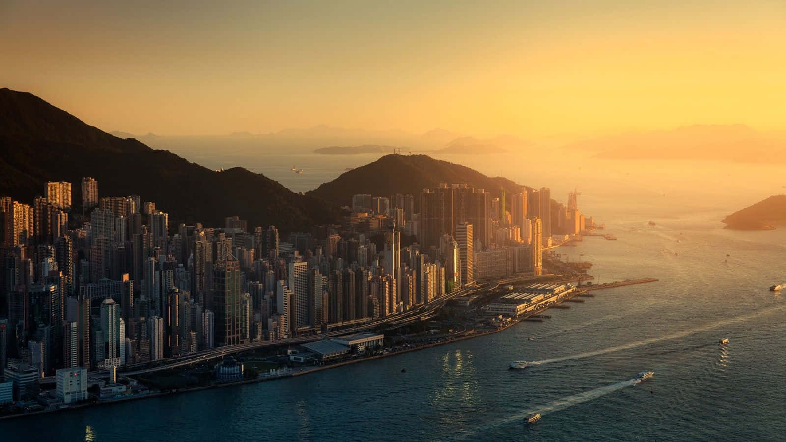 1600x900 桌布 壁紙 — 香港天際線在金色日落時的鳥瞰圖，摩天大樓在港口水面上反射溫暖的光線。