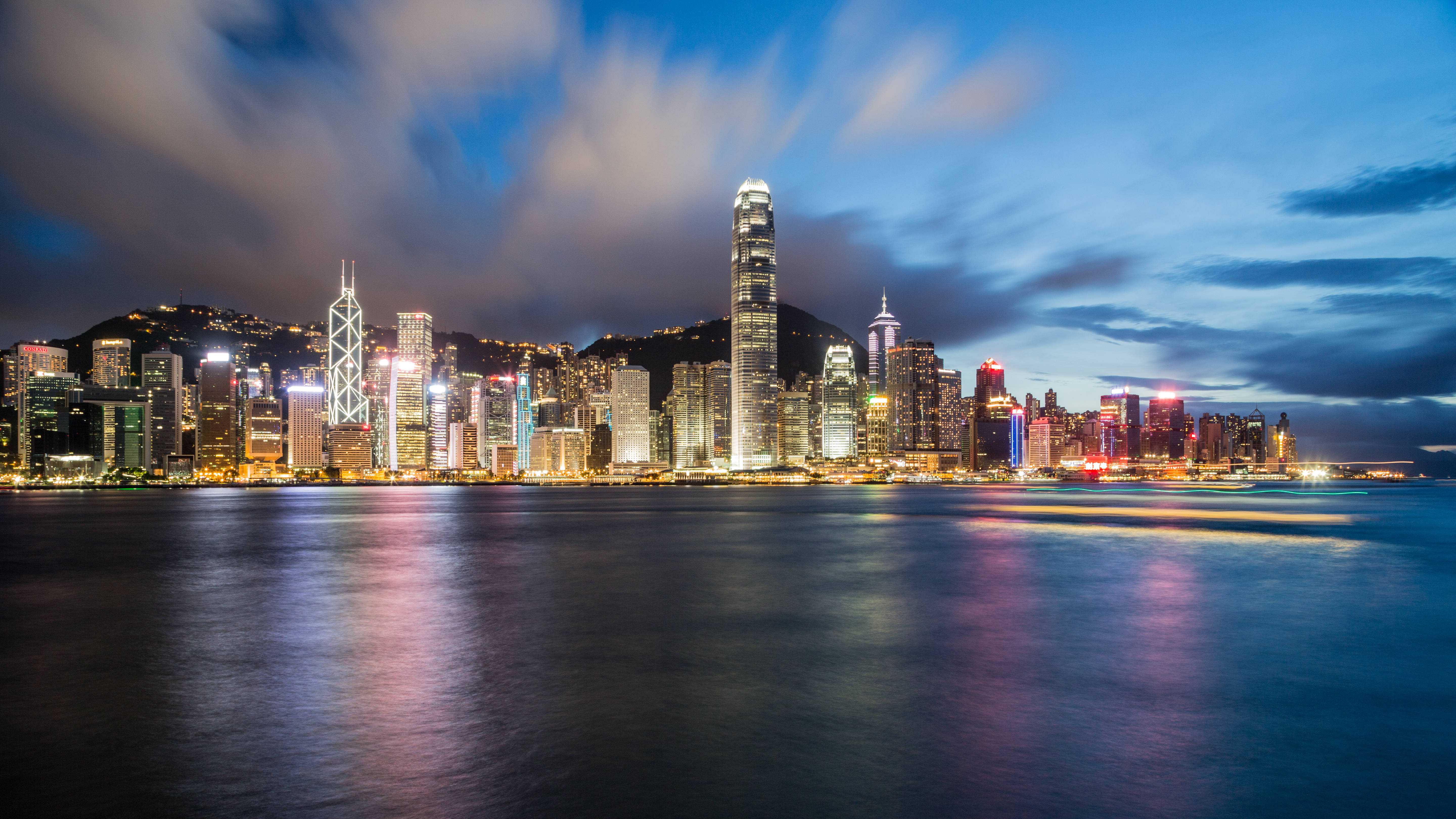 香港 桌布 壁紙 — 香港天際線在黃昏時分，模糊的雲層和發光的摩天大樓倒映在平靜的水面上。