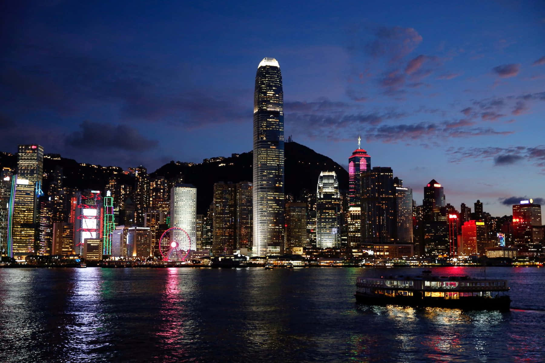 香港 桌布 壁紙 — 香港天際線在黃昏時分，燈火通明的摩天大樓和水上的渡輪反射出五彩繽紛的燈光。