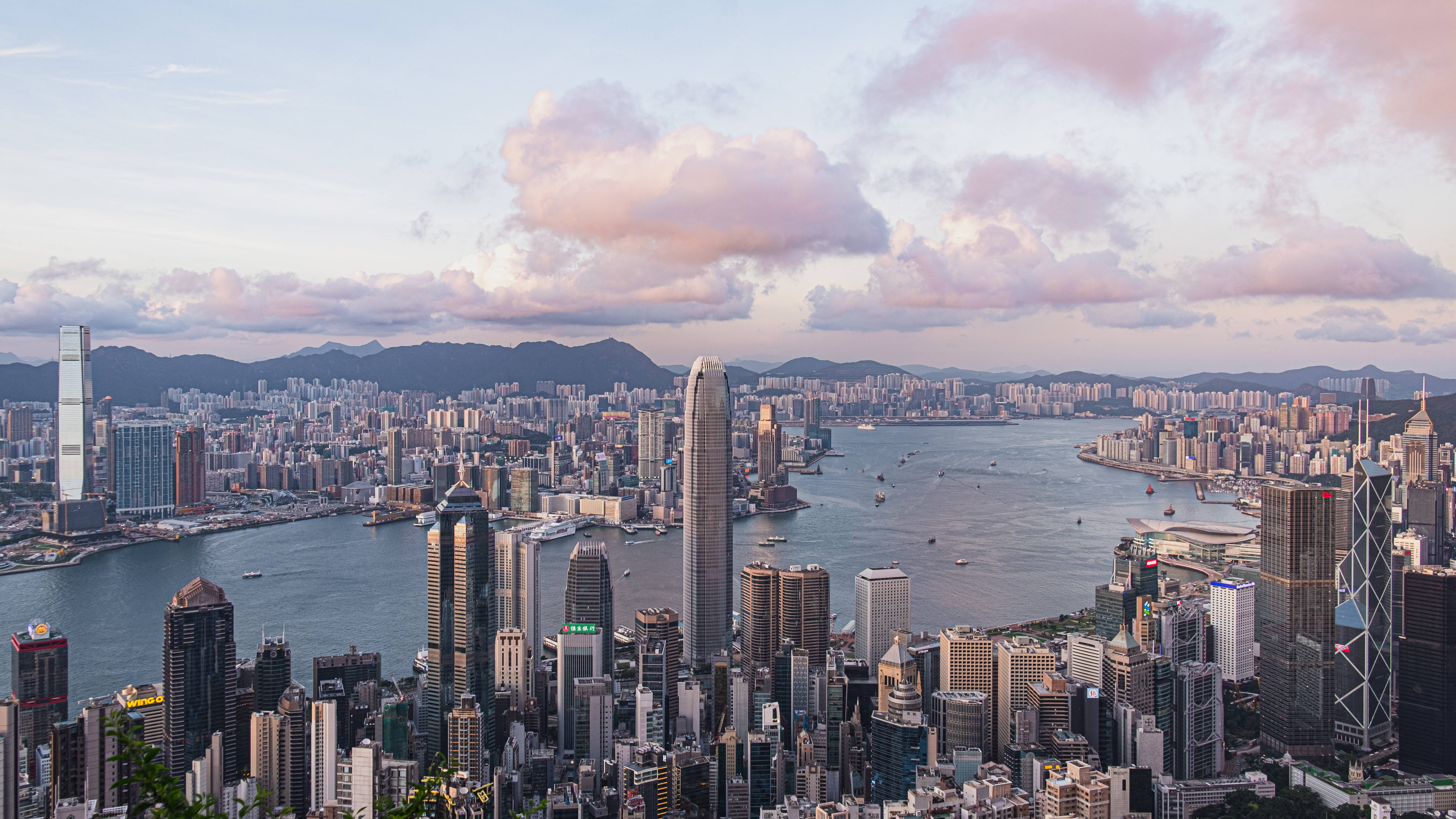 香港 桌布 壁紙 — 香港天際線的 panoramic 景觀，日落時分，粉紅色的雲朵在高聳的摩天大樓之上，海港中佈滿船隻。