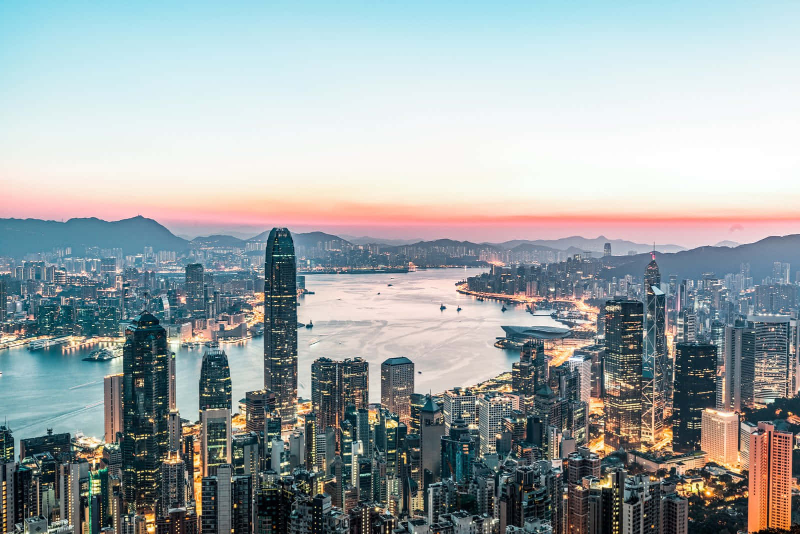 香港 桌布 壁紙 — 香港天際線的 panoramic 景觀，夕陽下閃爍的摩天大樓和寧靜的海港反射著粉紅色和藍色的天空。