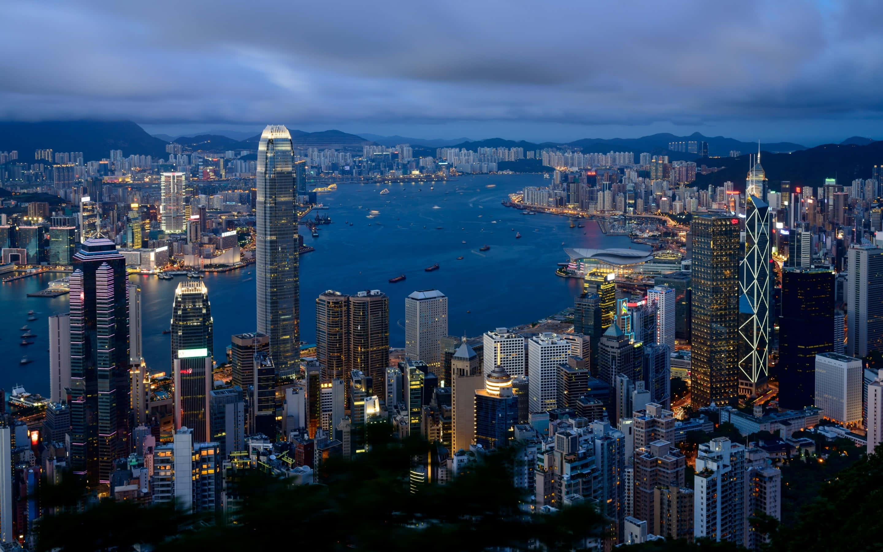 Macbook Retina 桌布 壁紙 — 香港天際線的 panoramic 黃昏景觀，發光的摩天大樓和點綴著船隻的港口，在陰沉天空下。
