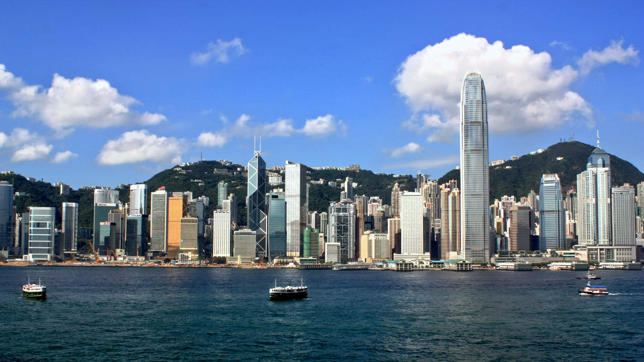香港 桌布 壁紙 — 香港天際線的全景視圖，高聳的摩天大樓和港口的船隻在明亮的藍天白雲下。