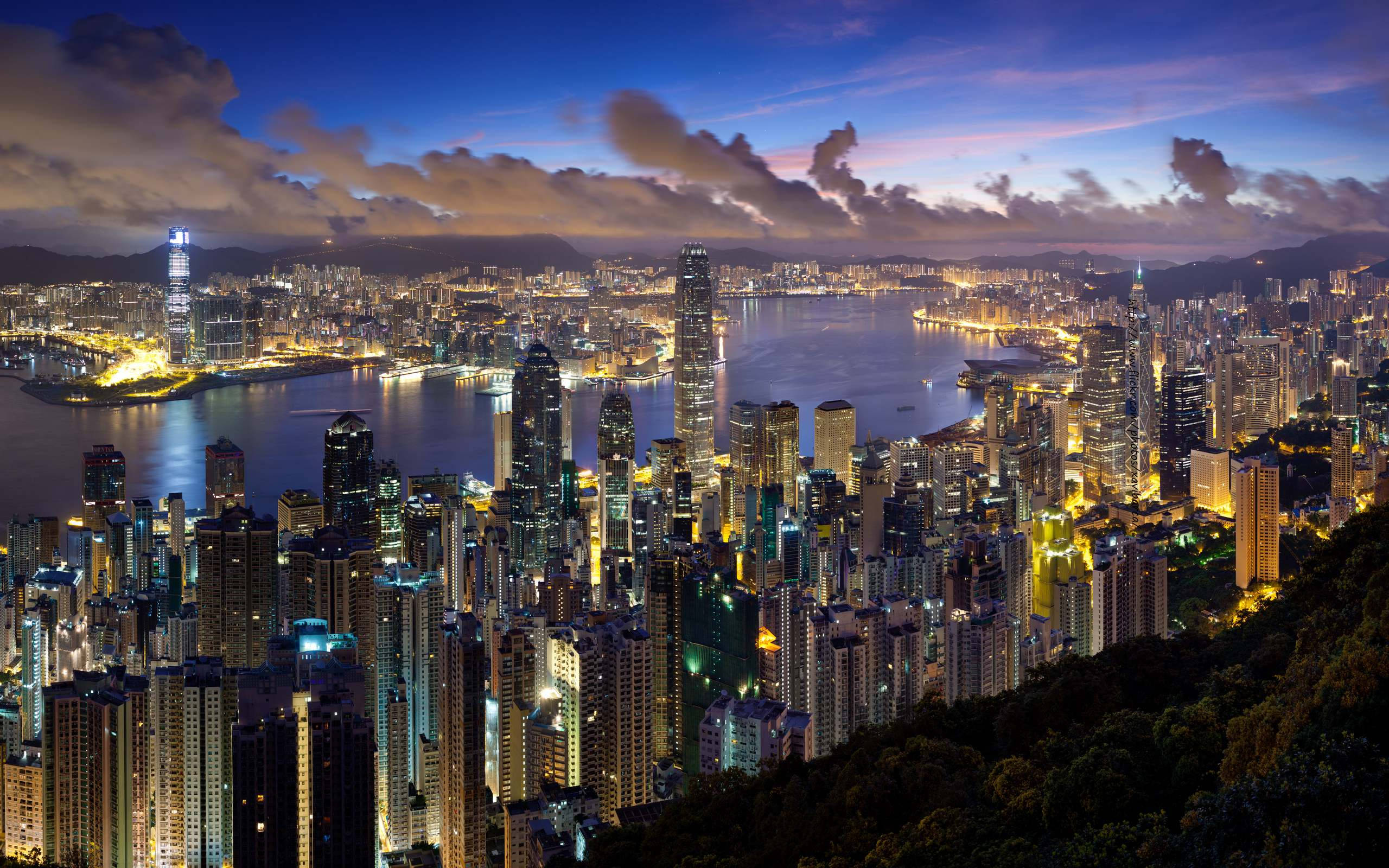雲朵 桌布 壁紙 — 香港天際線的壯麗暮色景觀，發光的摩天大樓在戲劇性的紫色和藍色天空下倒映在港口水域中。