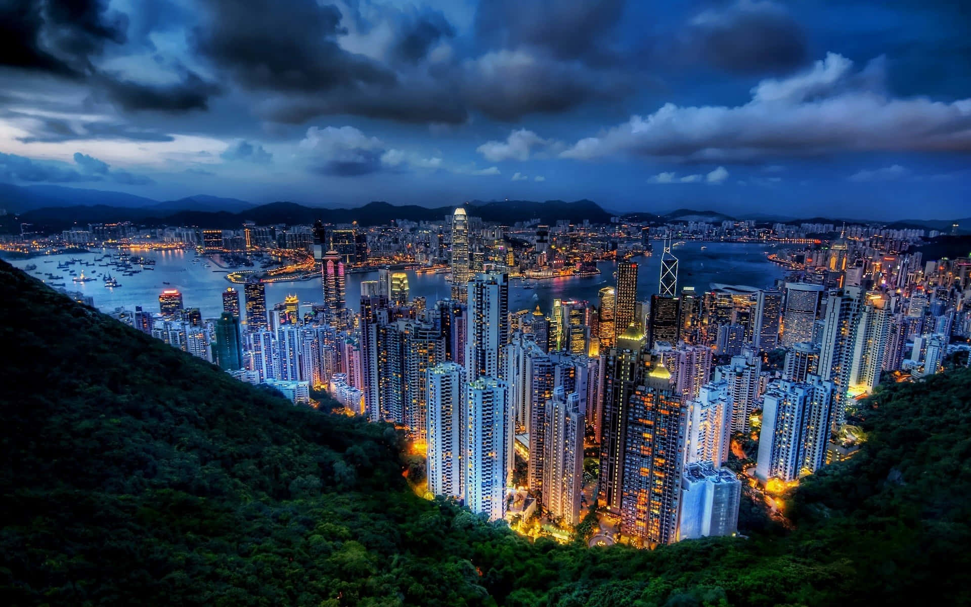 風景動畫 桌布 壁紙 — 香港天際線的壯麗黃昏景色，摩天大樓在鬱鬱蔥蔥的綠色山丘和點綴著船隻的港灣之間閃耀。