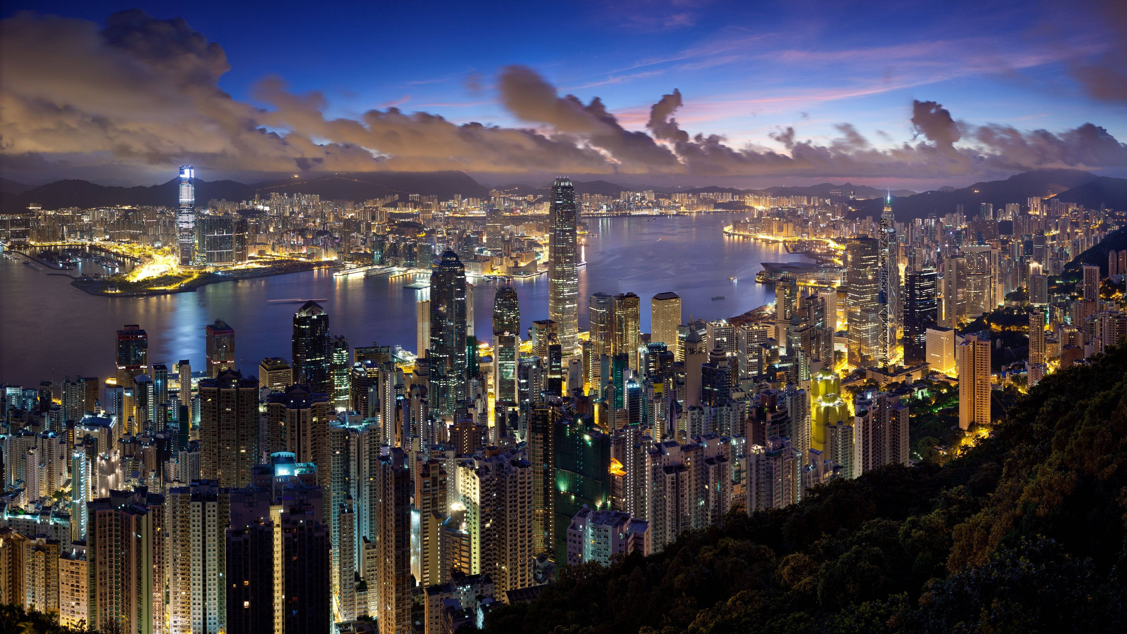 城市風景 桌布 壁紙 — 香港天際線的壯麗黃昏景色，發光的摩天大樓在戲劇性的紫色和藍色天空下倒映在港口水域中。