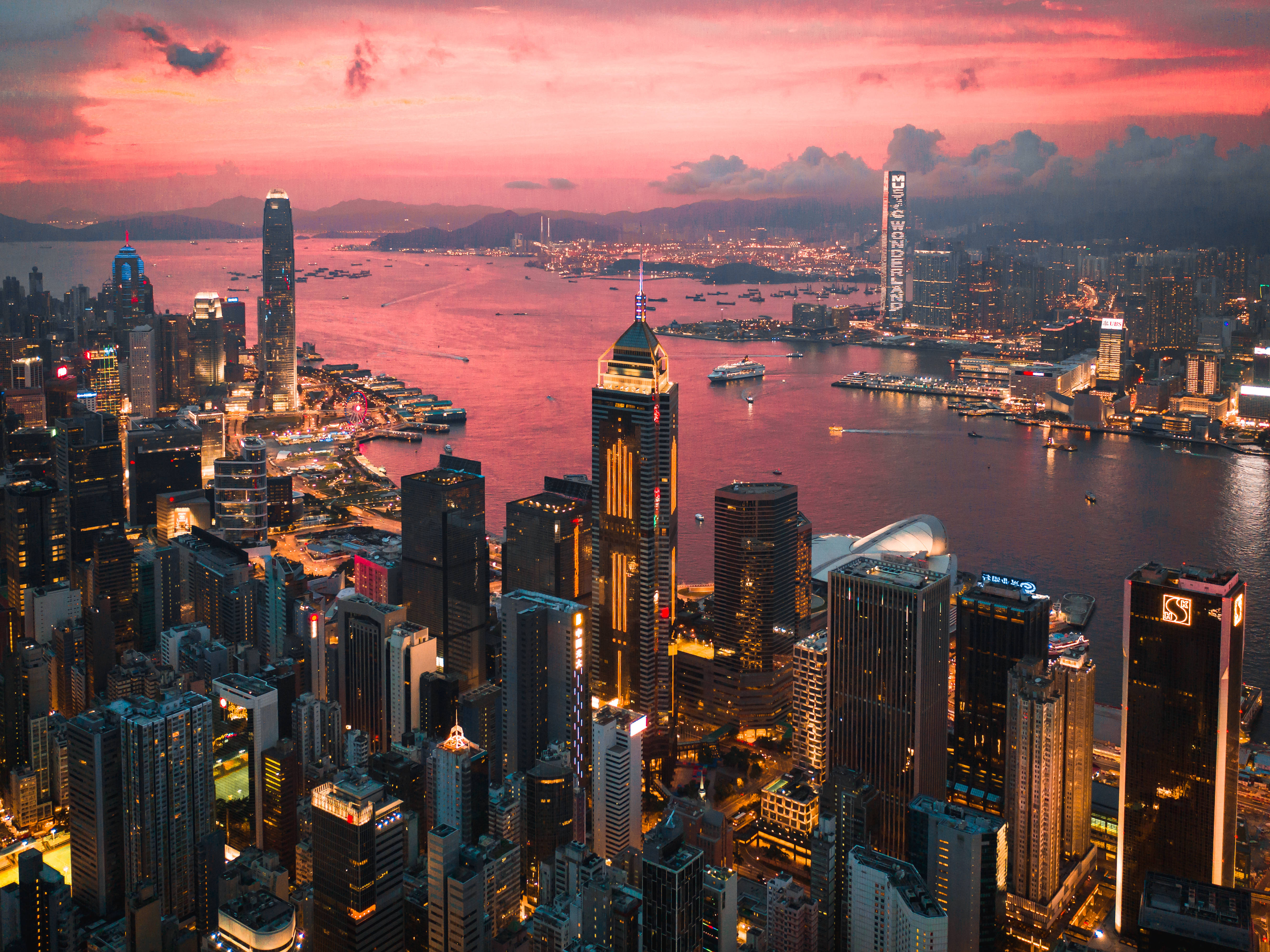 香港 桌布 壁紙 — 香港天際線的鳥瞰圖，日落時分，粉紅色的雲彩和發光的摩天大樓倒映在港灣水域中。