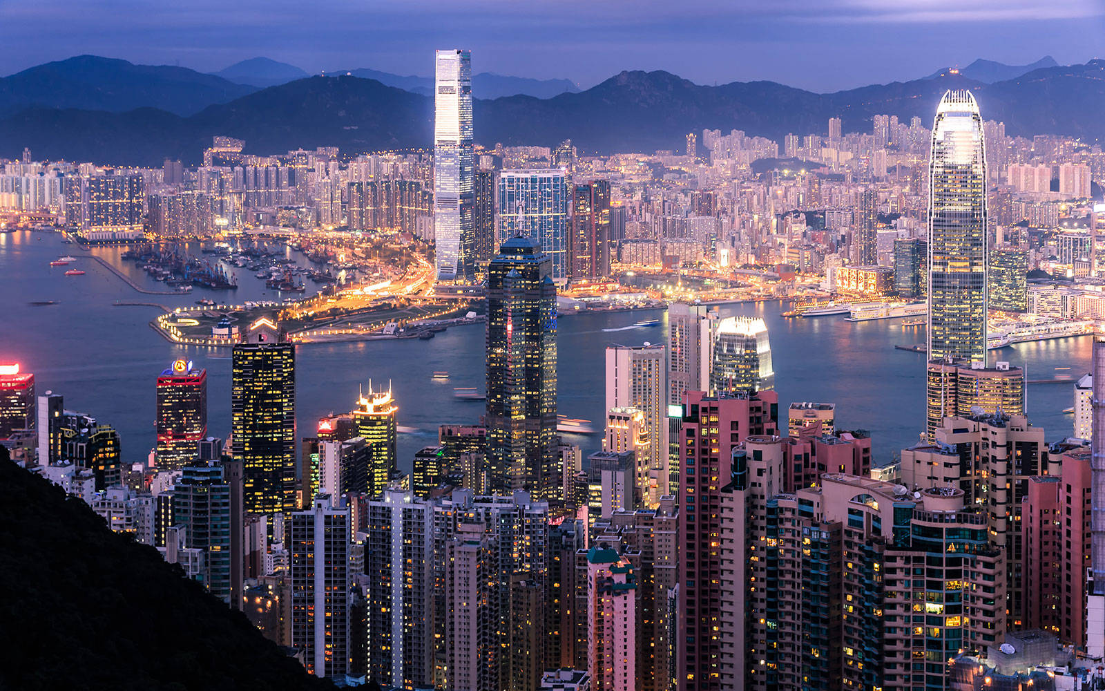 Asia 桌布 壁紙 — 香港天際線的黃昏全景，發光的摩天大樓和港口船隻映襯著黑暗的山脈。
