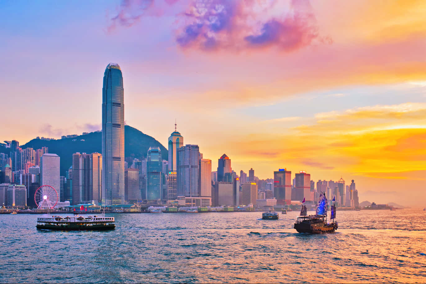 香港 桌布 壁紙 — 香港日落時分的天際線，充滿活力的橙色雲彩倒映在海港水面，船隻在高聳的摩天大樓旁邊滑過。