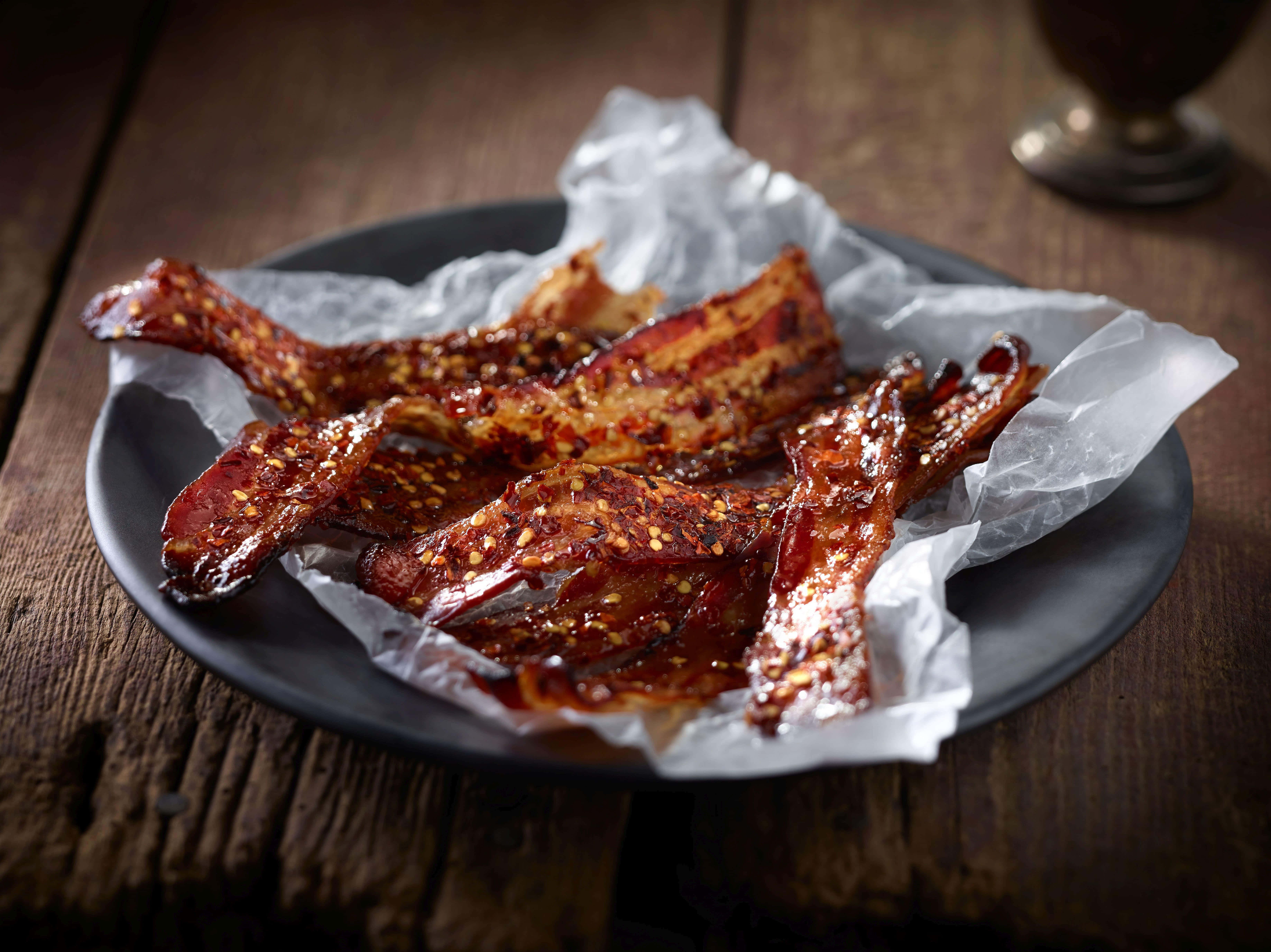 bacon 桌布 壁紙 — 香脆辣味培根條在烘焙紙上閃爍著醬汁和芝麻，放在深色盤子上，置於鄉村風木桌上。