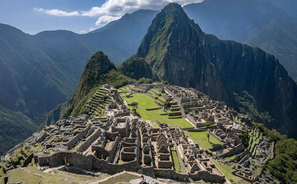 World Travel 桌布 壁紙 — 馬丘比丘的古老石頭遺址在陽光明媚的山脊上蔓延，周圍是茂密的綠色山峰，在明亮的藍天下。