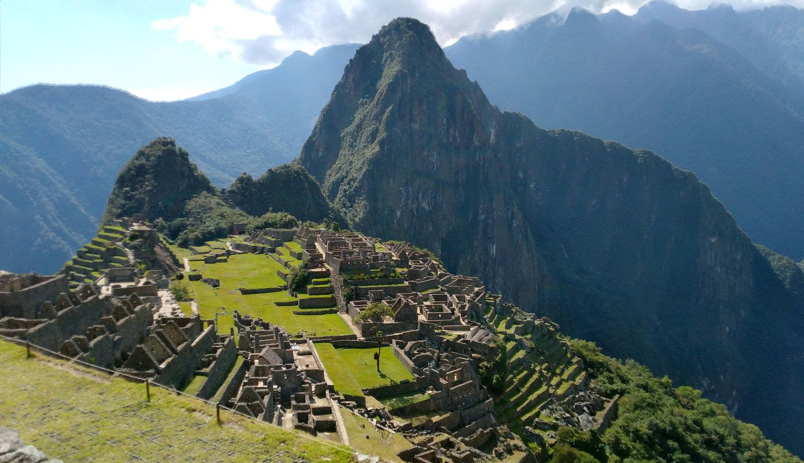 World Travel 桌布 壁紙 — 馬丘比丘的古老石頭遺址遍佈在陽光照耀的山脊上，背後是鬱鬱蔥蔥的綠色梯田和參差不齊的山峰。