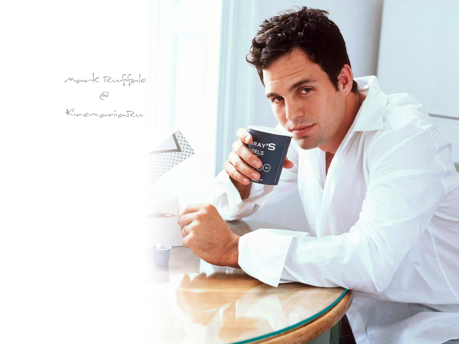 Mark Ruffalo 桌布 壁紙 — 馬克·盧法洛在明亮的極簡房間裡，在玻璃桌旁喝咖啡，柔和自然光灑落。