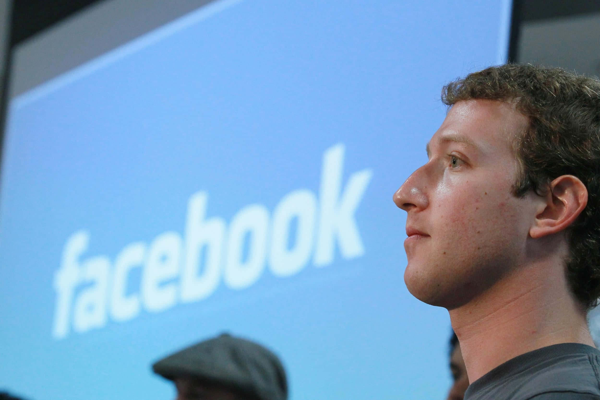 Facebook 桌布 壁紙 — 馬克·祖克柏側面站在一個顯示白色Facebook標誌的藍色大螢幕前。