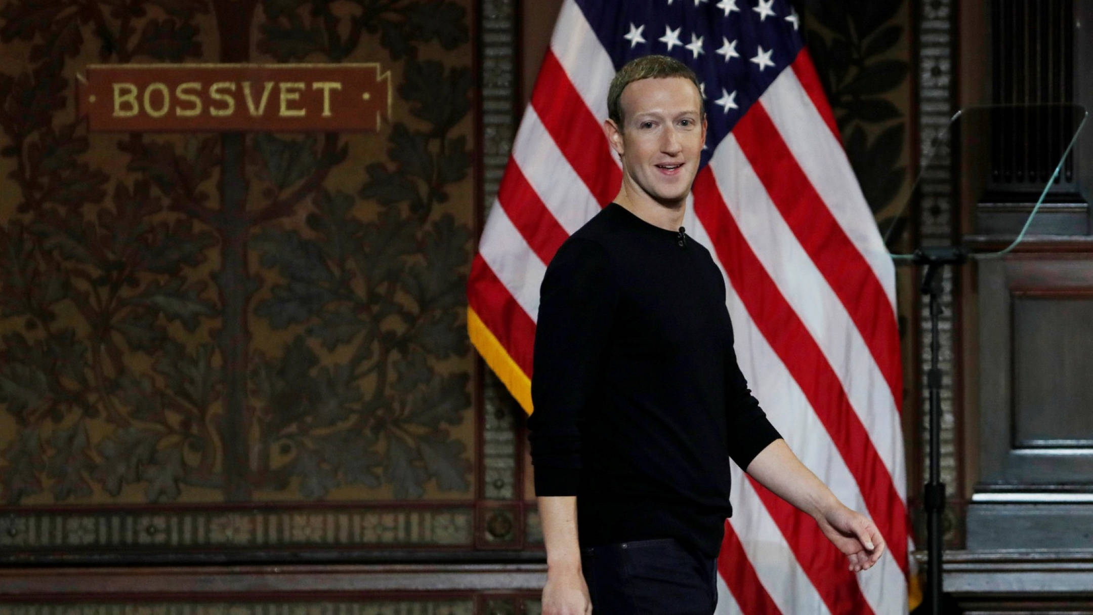 Zuckerberg 桌布 壁紙 — 馬克·祖克柏在正式房間內，站在美國國旗和「BOSSVET」標誌前微笑。