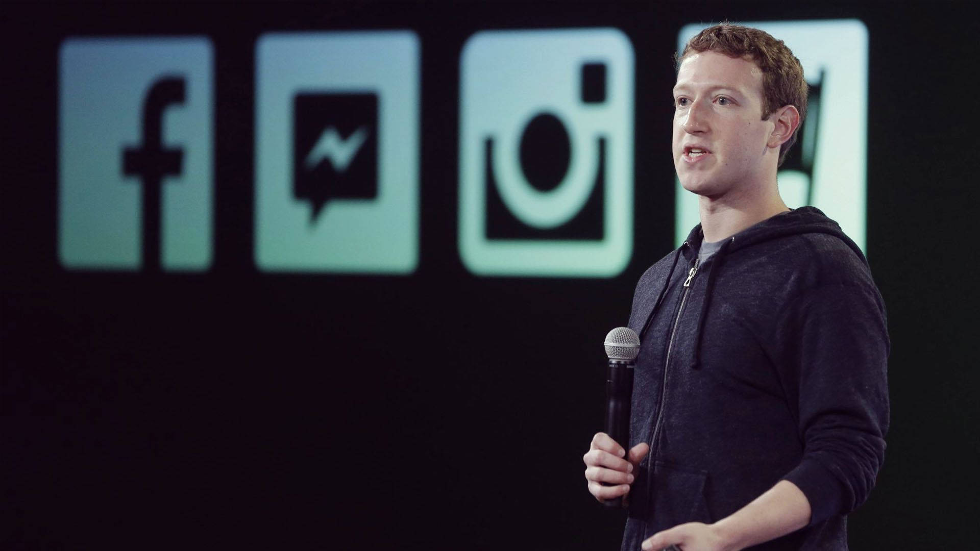 Zuckerberg 桌布 壁紙 — 馬克·祖克柏在舞台上對著麥克風講話，身後有發光的Facebook、Instagram和Messenger標誌。