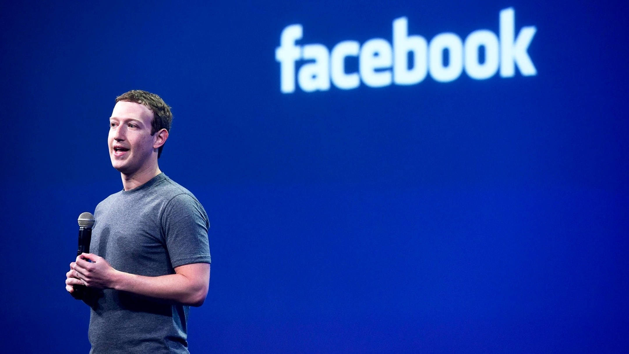 Zuckerberg 桌布 壁紙 — 馬克·祖克柏在舞台上對著麥克風講話，身後有一個大型藍色螢幕顯示Facebook標誌。