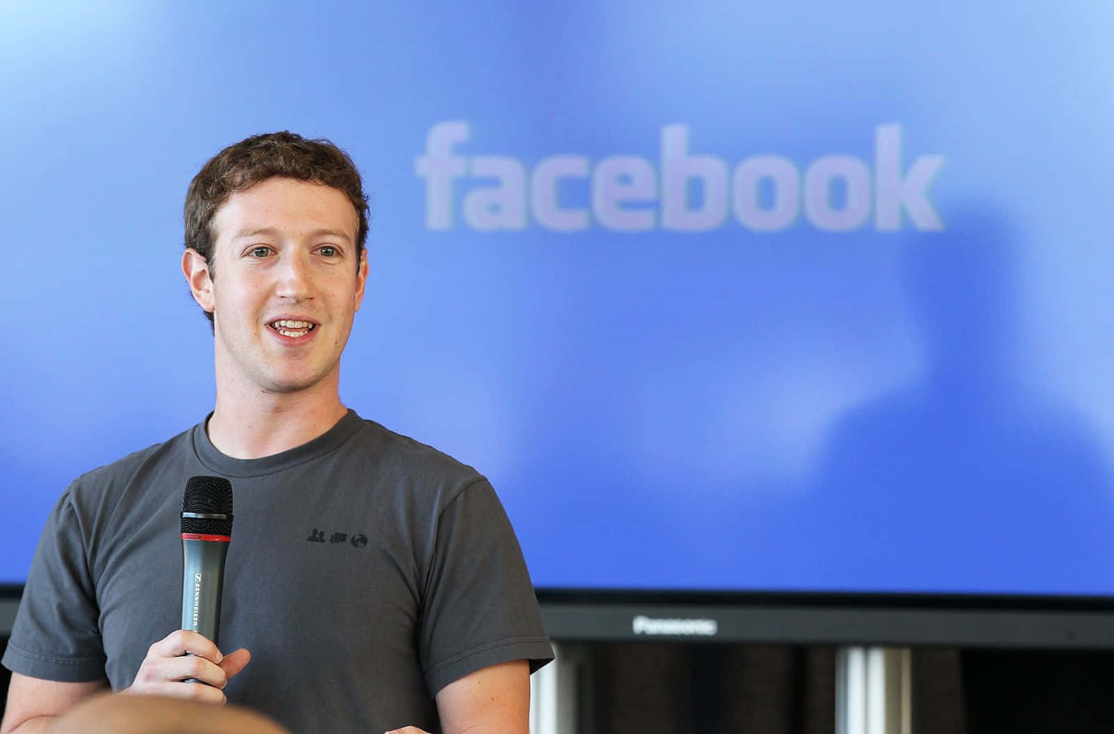 Facebook 桌布 壁紙 — 馬克·祖克柏在藍色Facebook標誌螢幕前微笑著拿著麥克風。