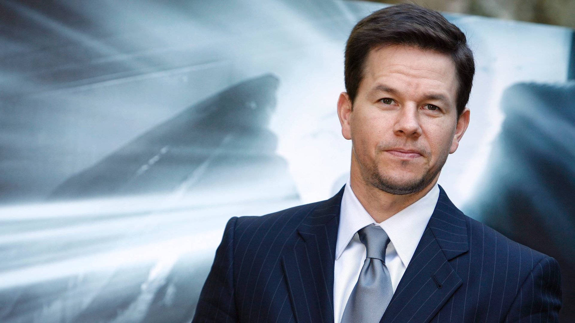 Mark Wahlberg 桌布 壁紙 — 馬克·華伯格穿著尖銳的細條紋西裝，自信地站在一輛高速行駛的汽車的模糊運動背景前。