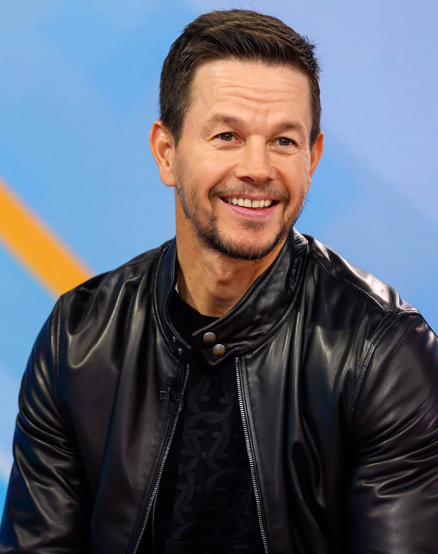 Mark Wahlberg 桌布 壁紙 — 馬克·華伯格穿著黑色皮夾克，在藍色和橙色背景前溫暖地微笑。