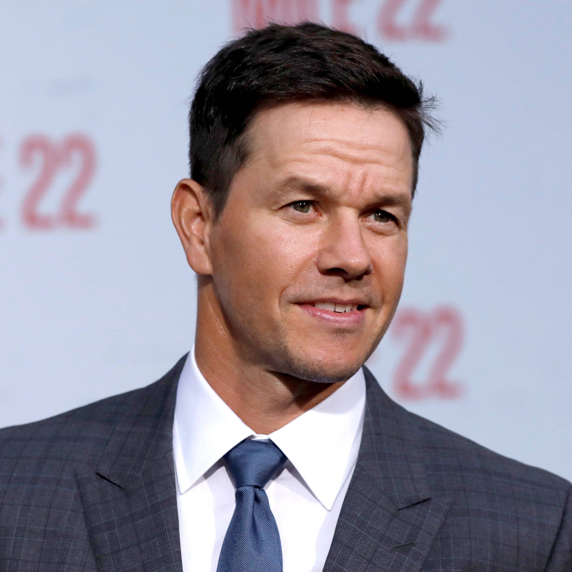 Mark Wahlberg 桌布 壁紙 — 馬克·華伯格身穿俐落灰色西裝和藍色領帶，在模糊的「22」背景的紅地毯活動上微妙地微笑。