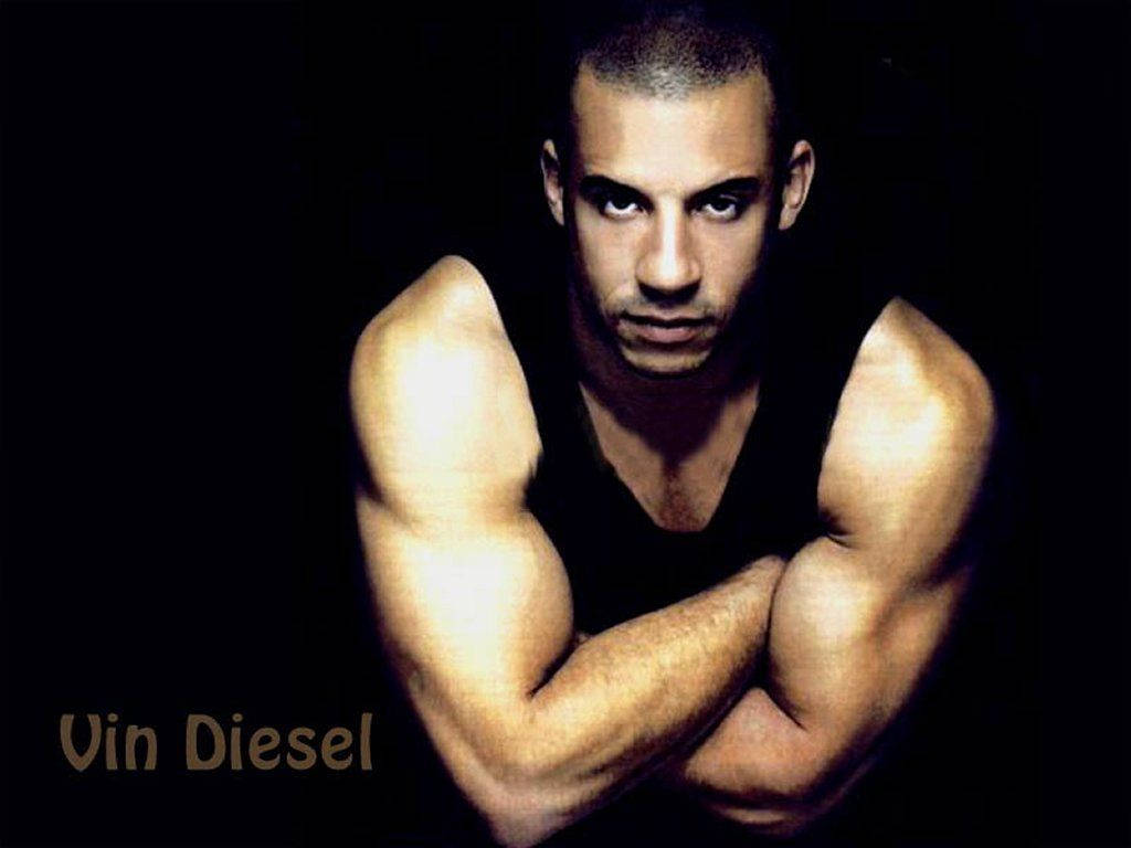 Diesel 桌布 壁紙 — 馮·迪索雙臂交叉，在黑色背景前凝視，戲劇性燈光突顯其肌肉發達的身形。
