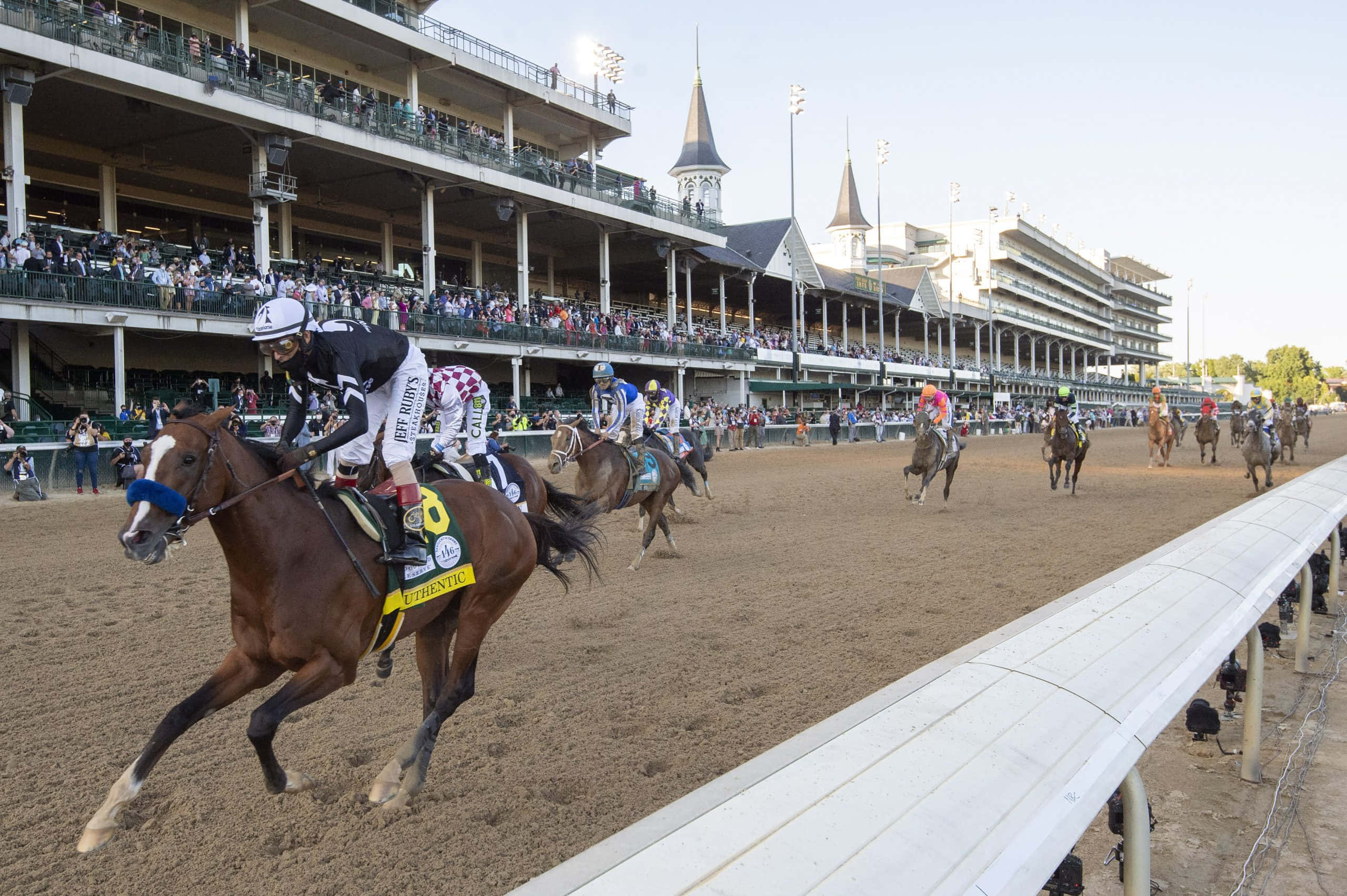 Kentucky Derby 桌布 壁紙 — 騎師在泥地賽道上策騎純種馬競賽，觀眾在晴朗天空下擠滿看台。
